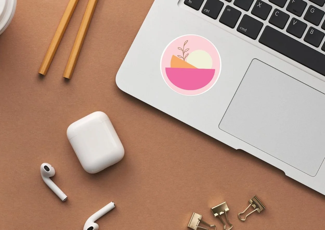 Top view of a workspace with a silver laptop displaying a pink, orange, and white abstract sticker, nearby a pair of white wireless earbuds with a charging case, some gold binder clips, a pair of wooden chopsticks, and a small part of a cup on a brown surface.