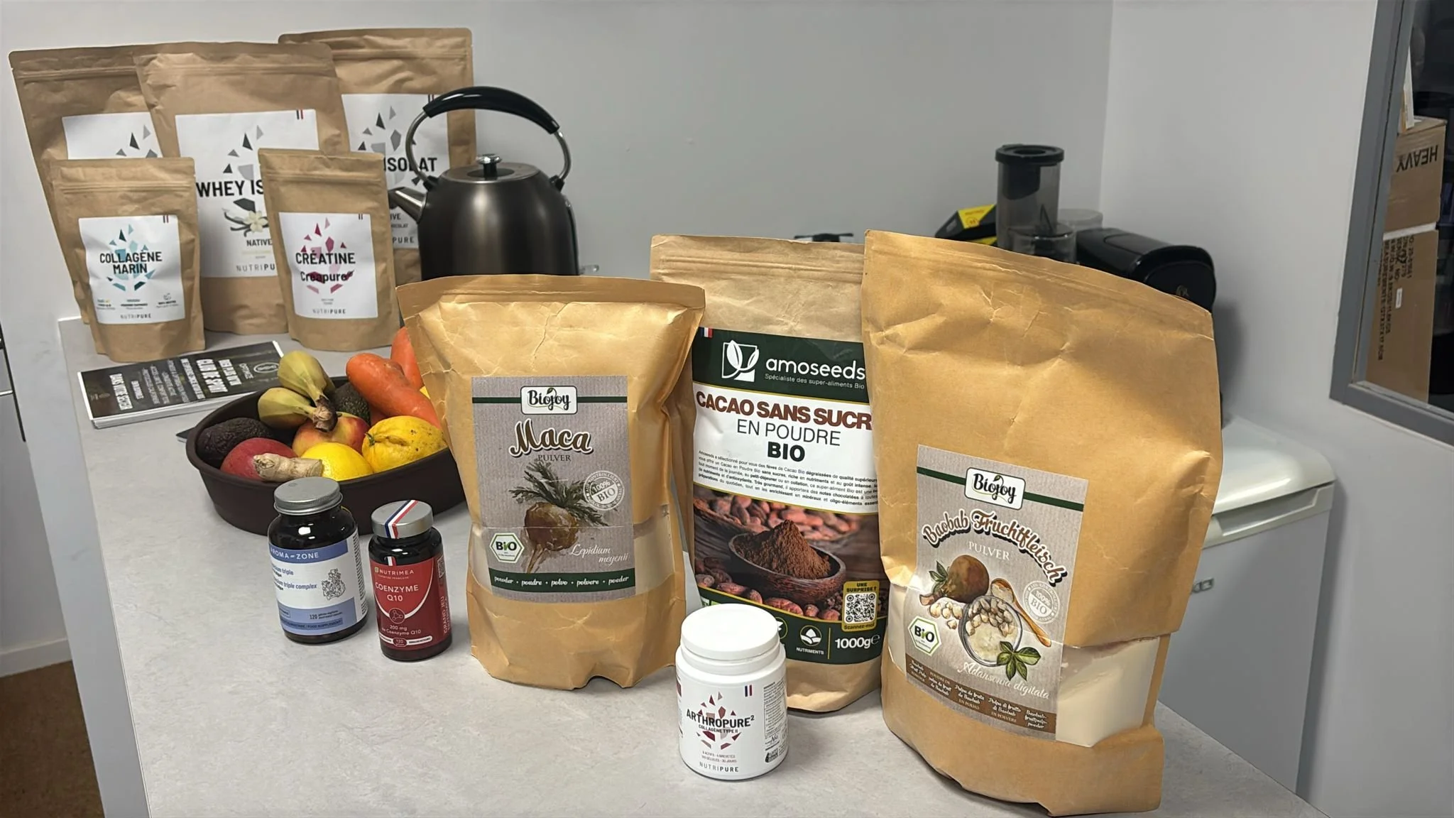 Produits naturels en poudre et compléments alimentaires sur un comptoir, fruit frais, bouilloire, et appareils de cuisine.