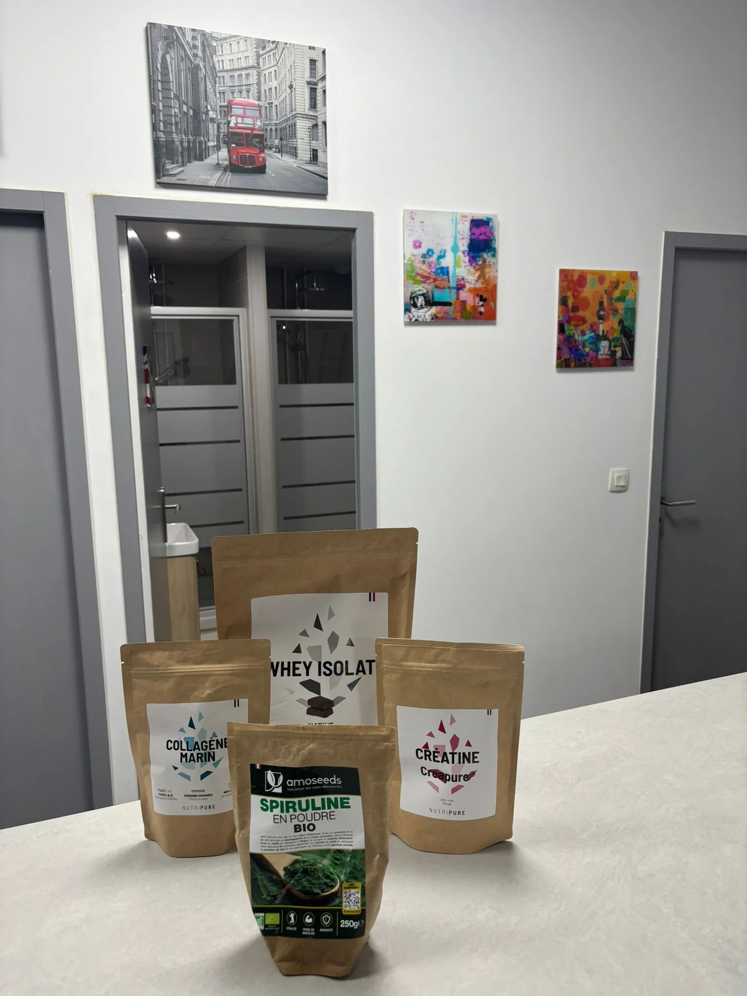 Divers compléments alimentaires bio et en poudre sur un comptoir dans une pièce avec des œuvres d'art colorées sur les murs et deux portes en arrière-plan.