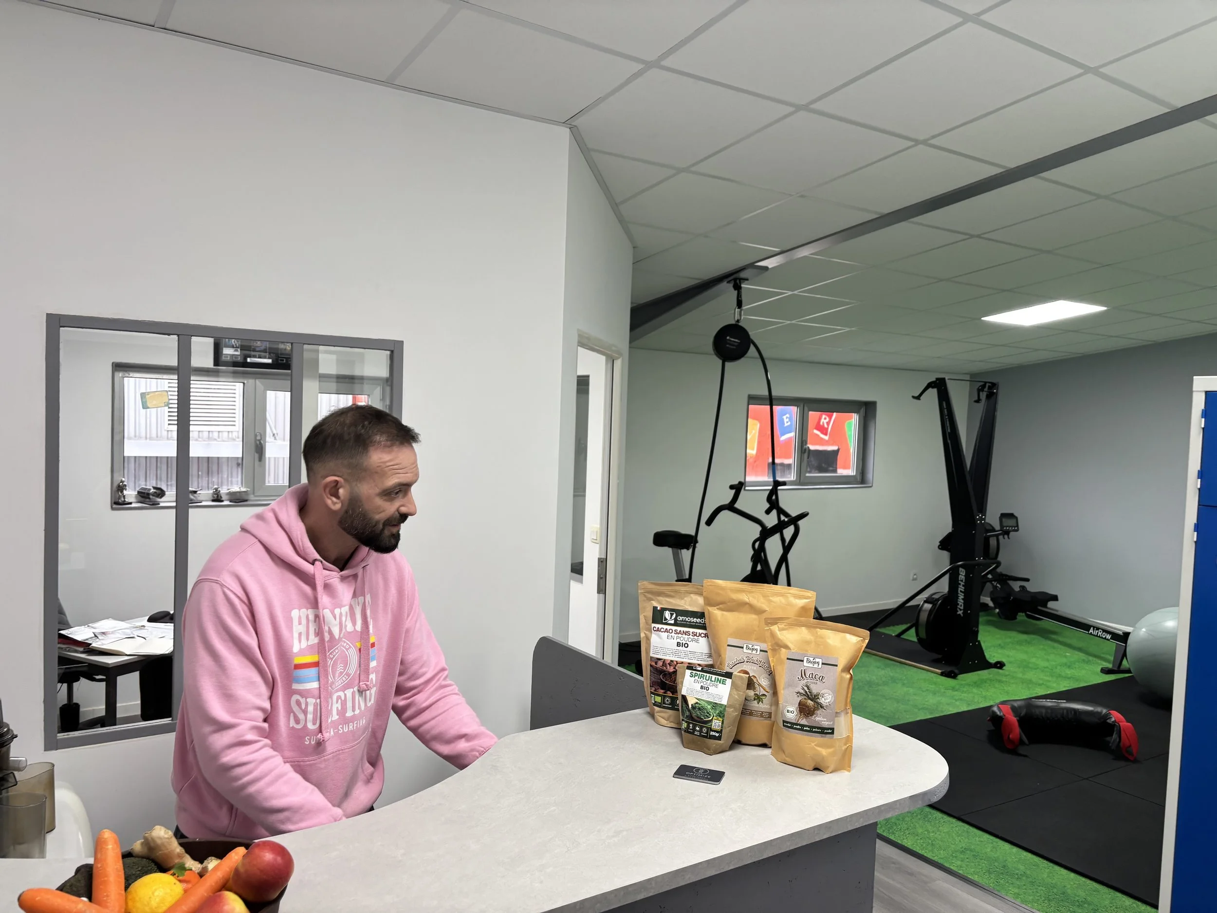 Un homme en pull rose est assis à un comptoir dans une salle de sport, avec des sacs de produits bio sur le comptoir. En arrière-plan, on voit des équipements de fitness comme un vélo d'appartement, un rameur, une balle d'exercice et un sac de frappe