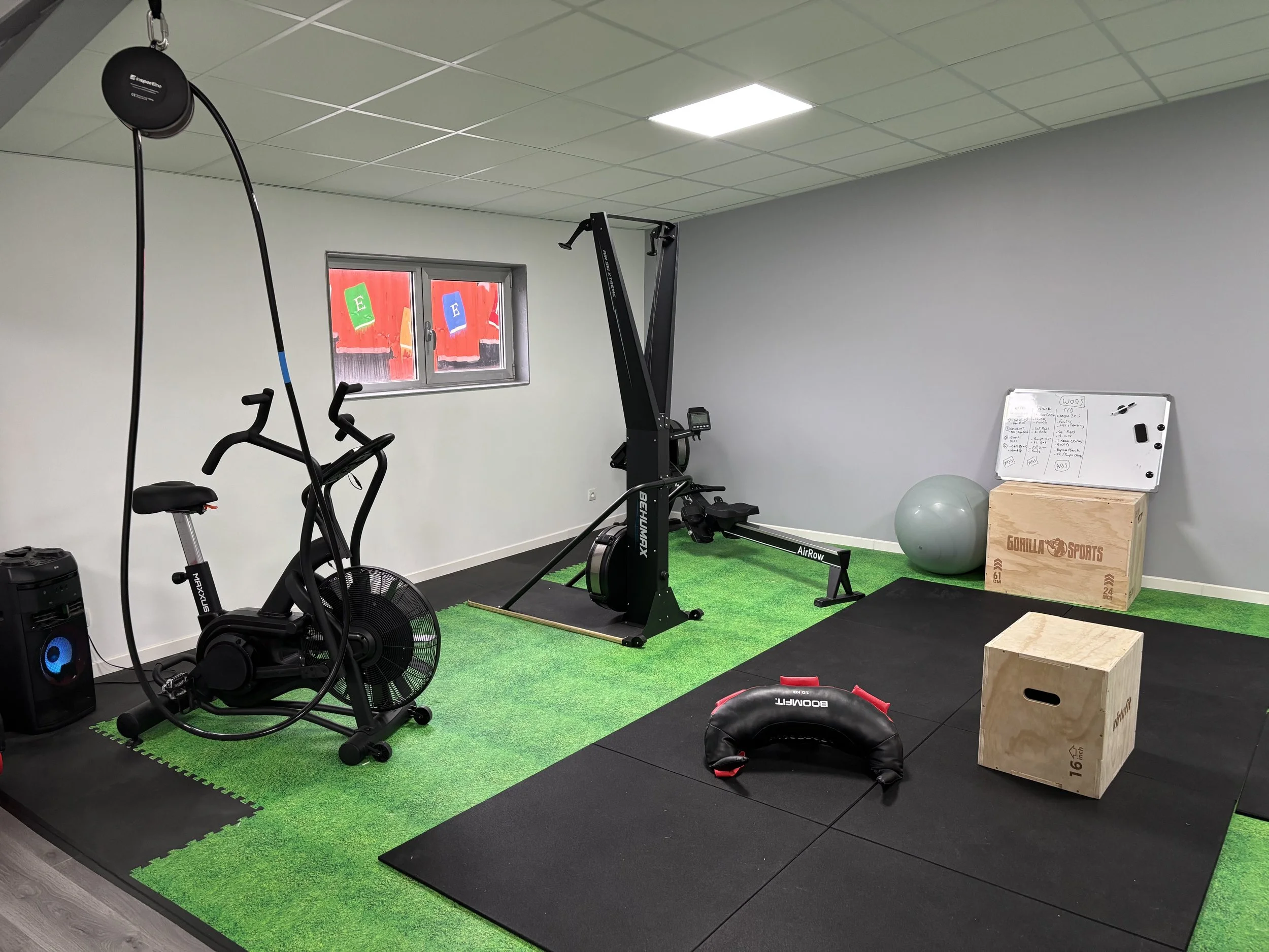 Salle de sport avec un vélo elliptique, un rameur, une balle de stabilité, une boîte en bois, une corde à sauter, un tableau blanc, un ballon de médecine, et un tapis d'entraînement.