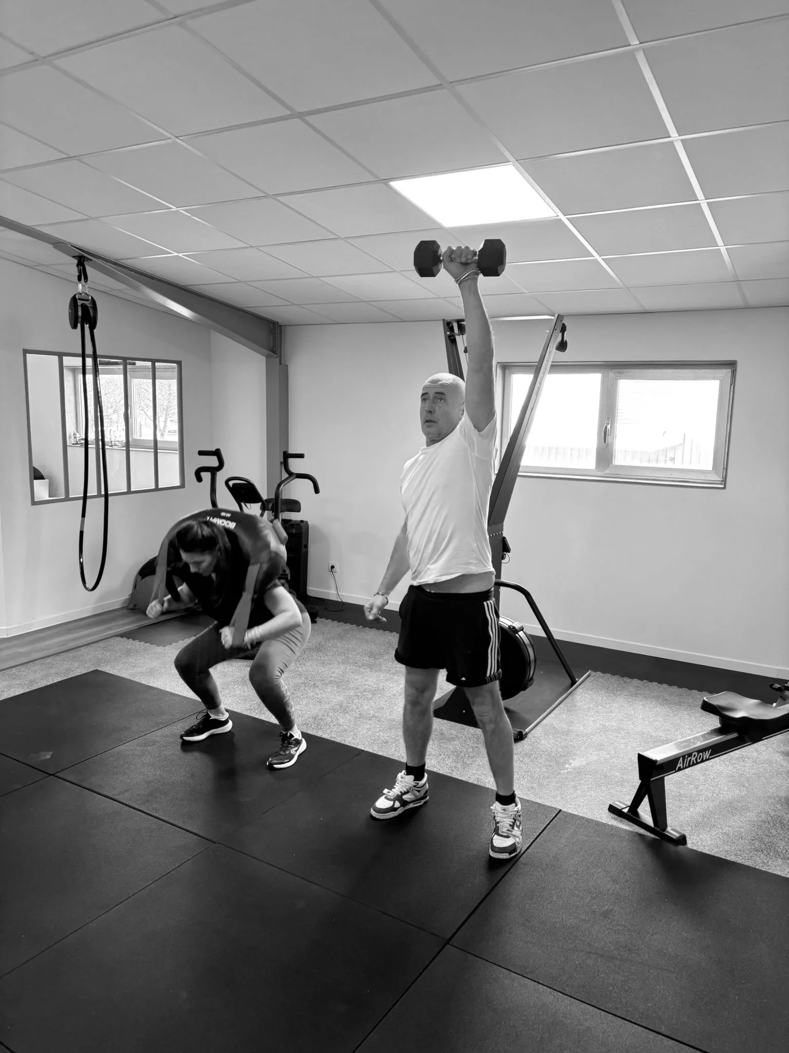 Deux personnes entraînées dans une salle de sport, un homme soulève un haltère avec le bras tendu vers le haut, une femme fait un exercice de squat ou de bent-over row, le tout en noir et blanc.