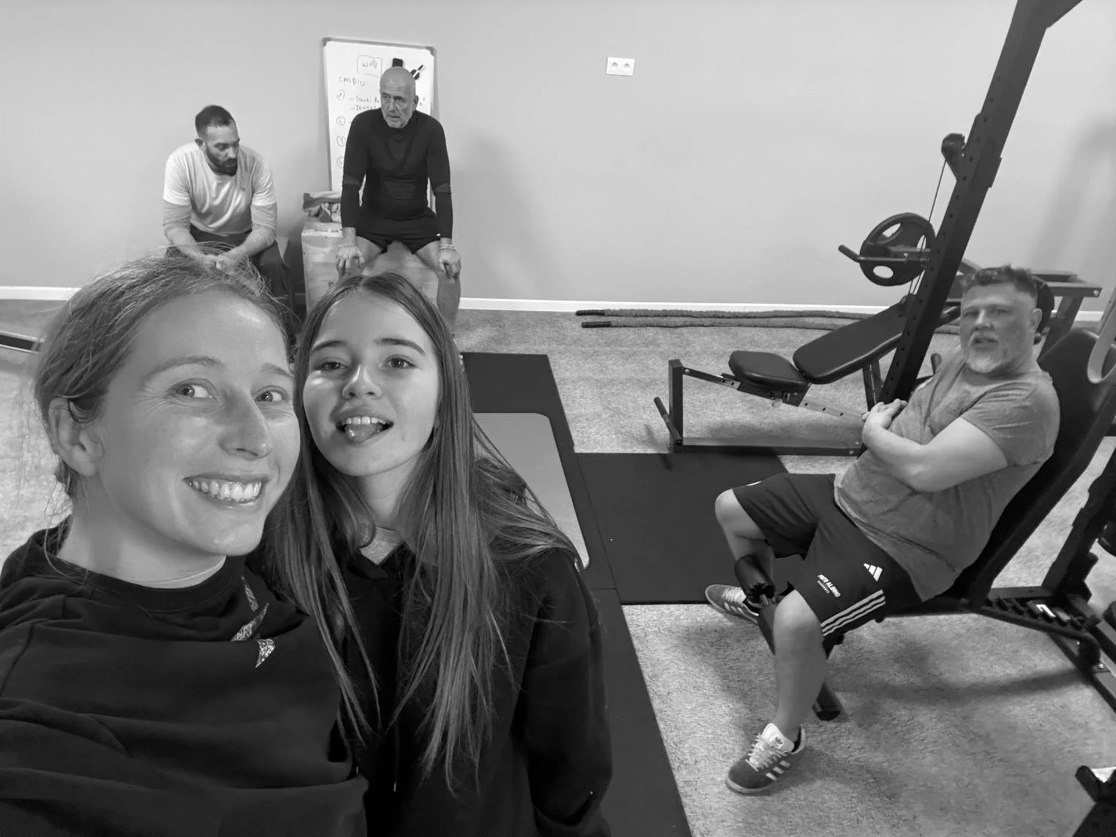 Cinq personnes dans une salle de sport, deux en premier plan prenant un selfie, trois à l'arrière, deux assises et une sur un banc, en train de faire de l'exercice.