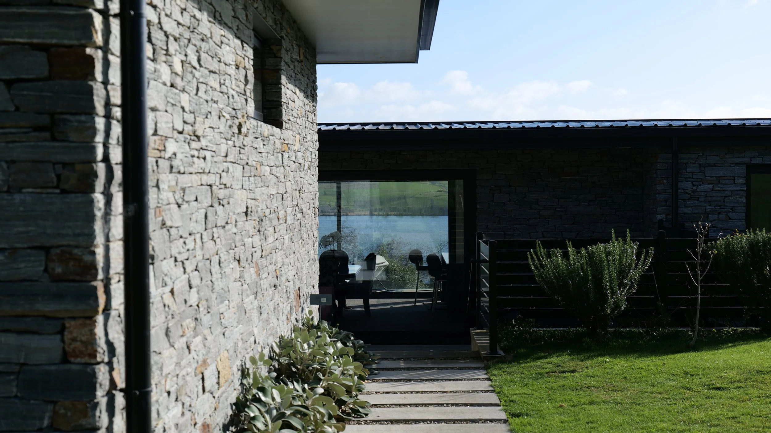 View of a modern house with stone exterior walls, a patio area with chairs and a table, and a scenic lake view in the background.