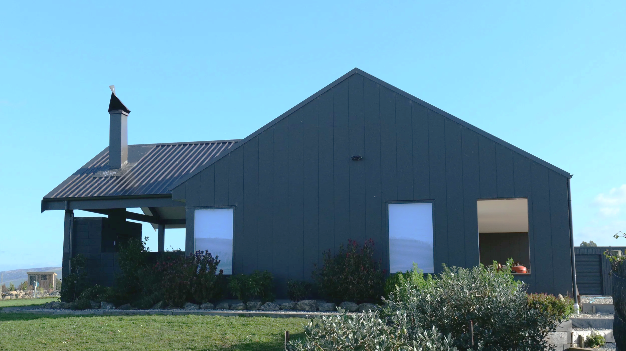 A modern house with dark gray exterior walls, a metal roof, and a chimney, under a clear blue sky. The house has large windows and a small garden in front.