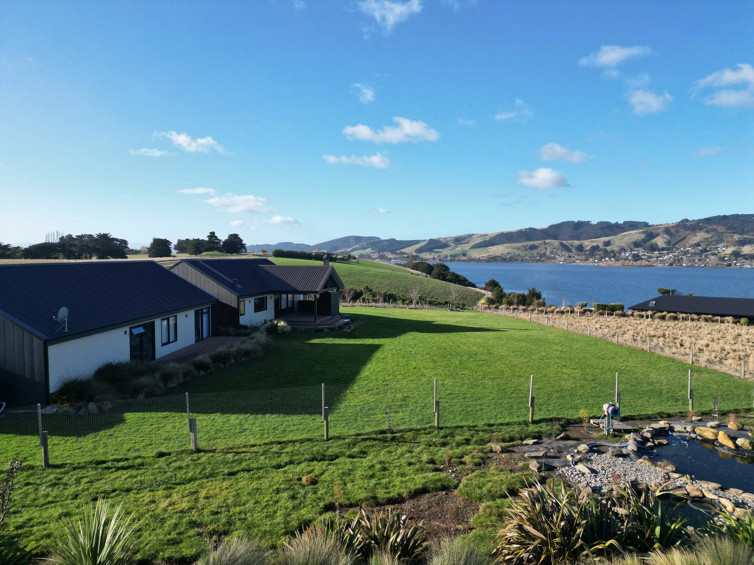 A scenic view of a lake with mountains in the background, green hills, and a modern house with a metal roof in the foreground.