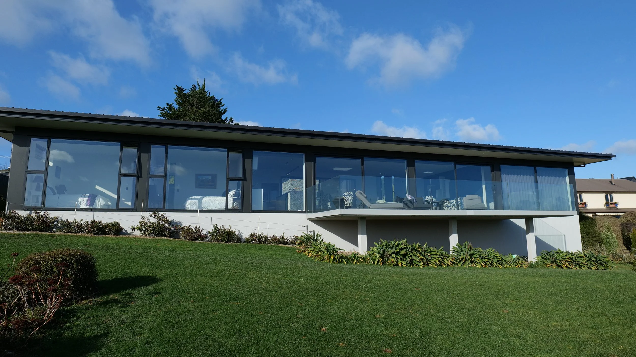 Modern house with large glass windows, a glass-enclosed balcony, and a well-maintained front yard with green grass and bushes under a blue sky with scattered clouds.