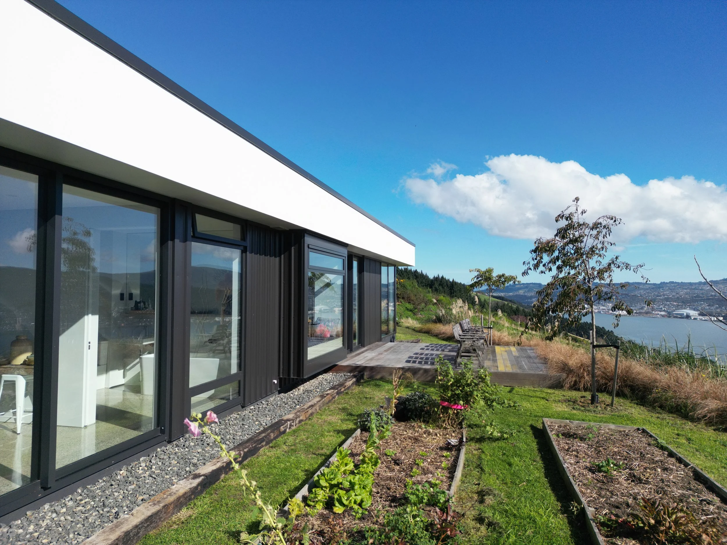 Modern house with large windows on a hillside overlooking a bay, with a garden and benches outside under a clear blue sky.