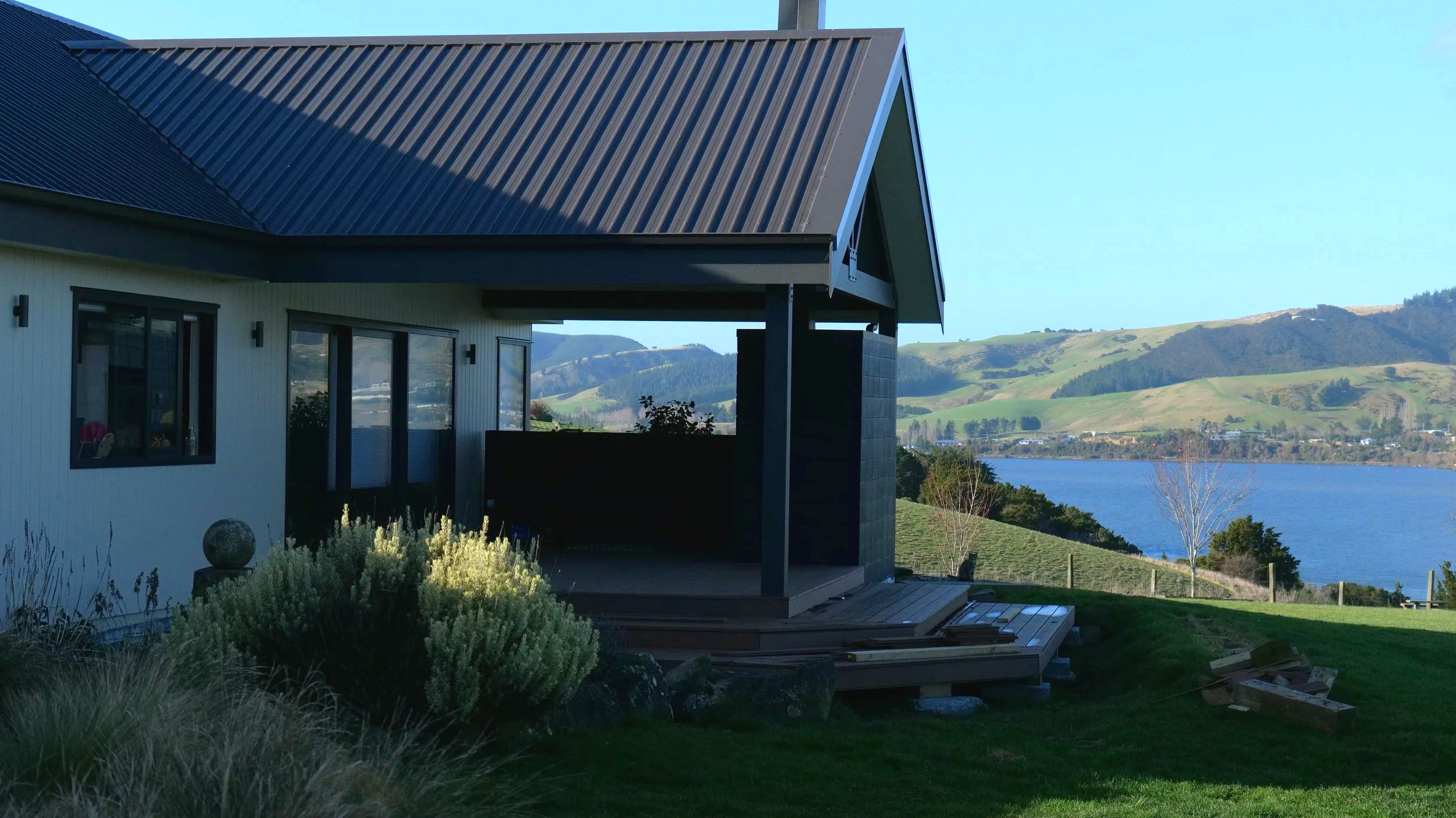 View of a house with a black metal roof, white siding, large windows, and a deck overlooking a lake and green rolling hills in the background.