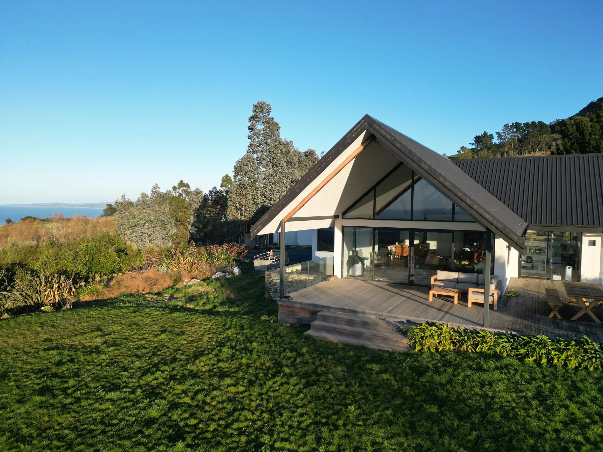 Modern house with large glass windows and a sloped roof in a green landscape with trees and mountains in the background, clear blue sky overhead.
