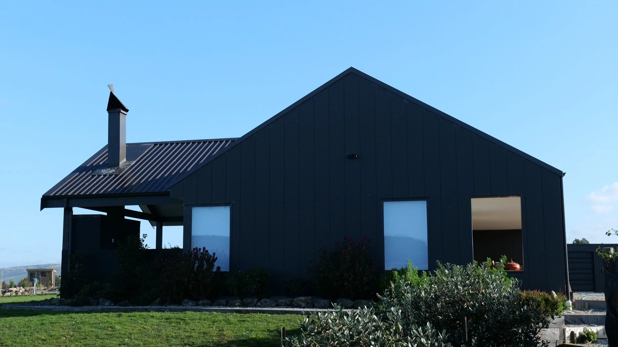 A modern dark gray house with a sharply peaked roof, a chimney, and several small windows, set against a clear blue sky.