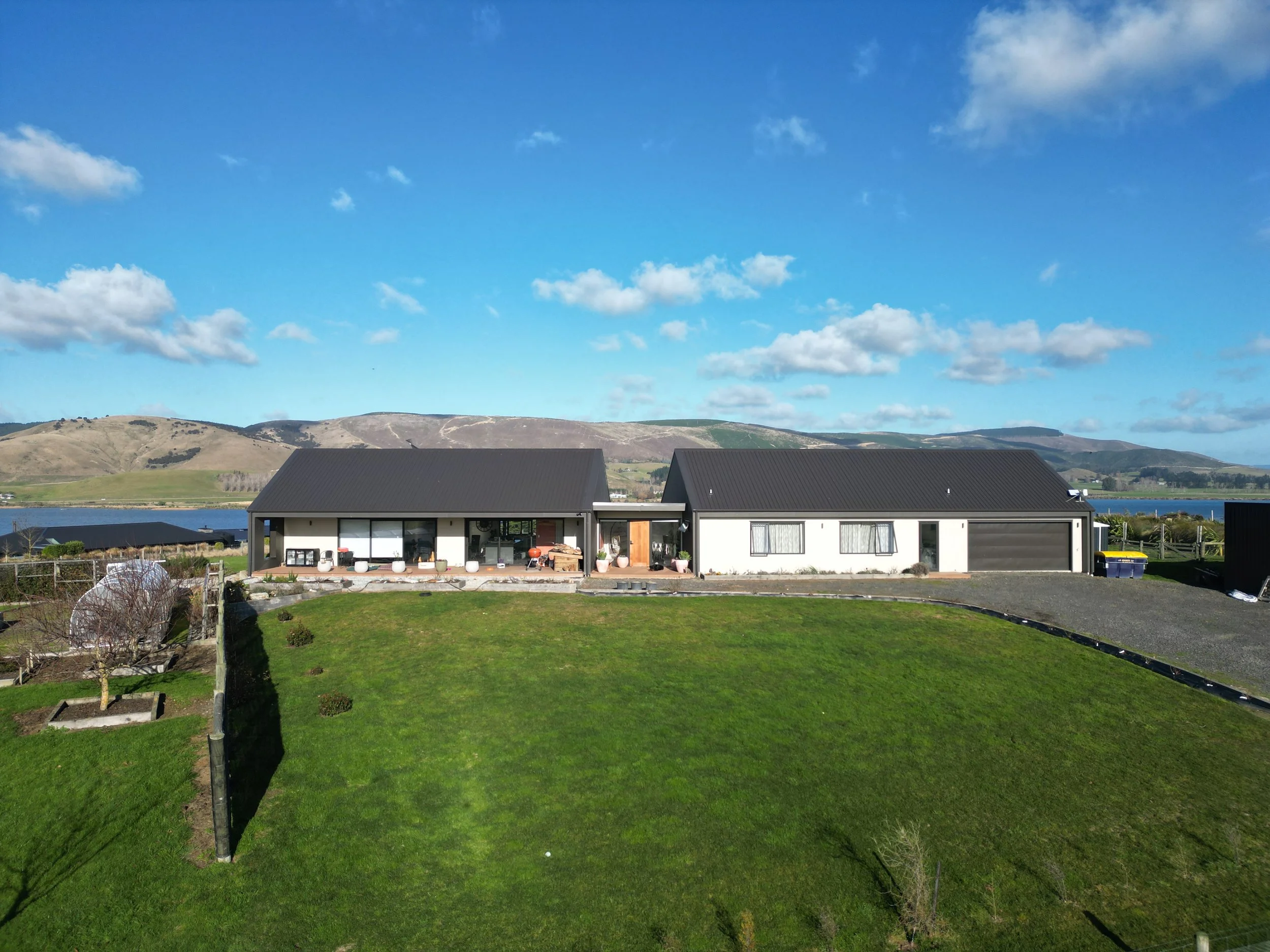 A backyard with a well-maintained green lawn, garden beds, and a modern house with a black roof and white walls. In the background, there are rolling hills and a body of water, under a blue sky with scattered clouds.