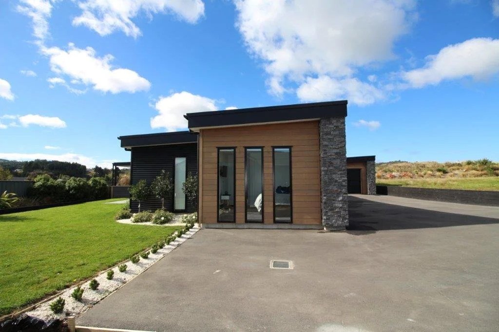 Modern house with a combination of wood, stone, and dark siding, large windows, and a paved driveway on a sunny day with blue sky and clouds.