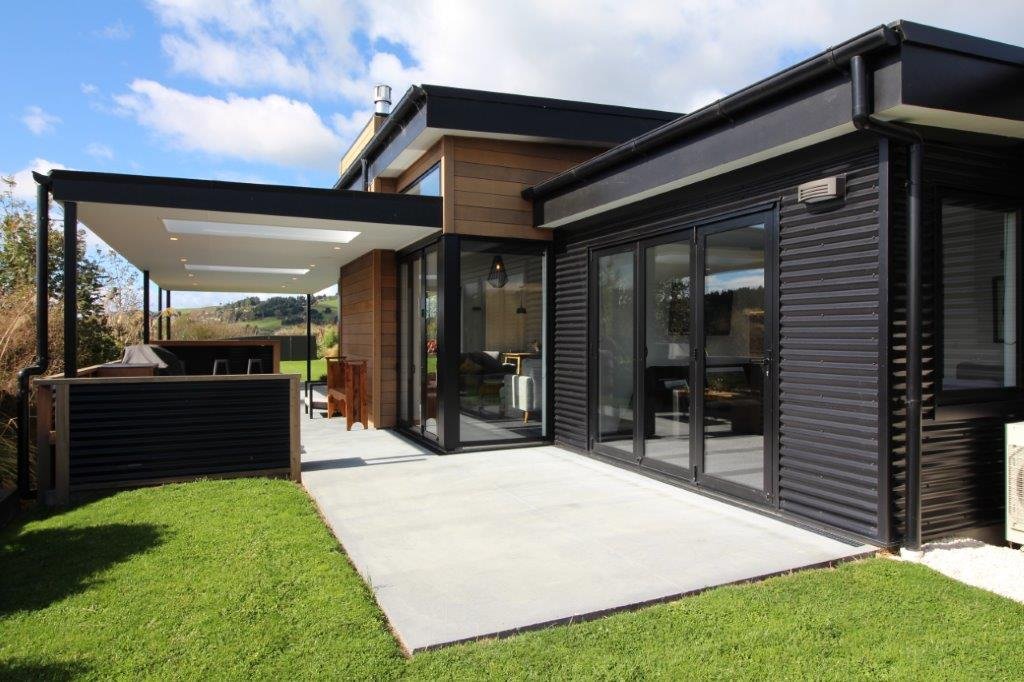 Modern house with black siding, large sliding glass doors, a concrete patio, and a lawn with green grass.