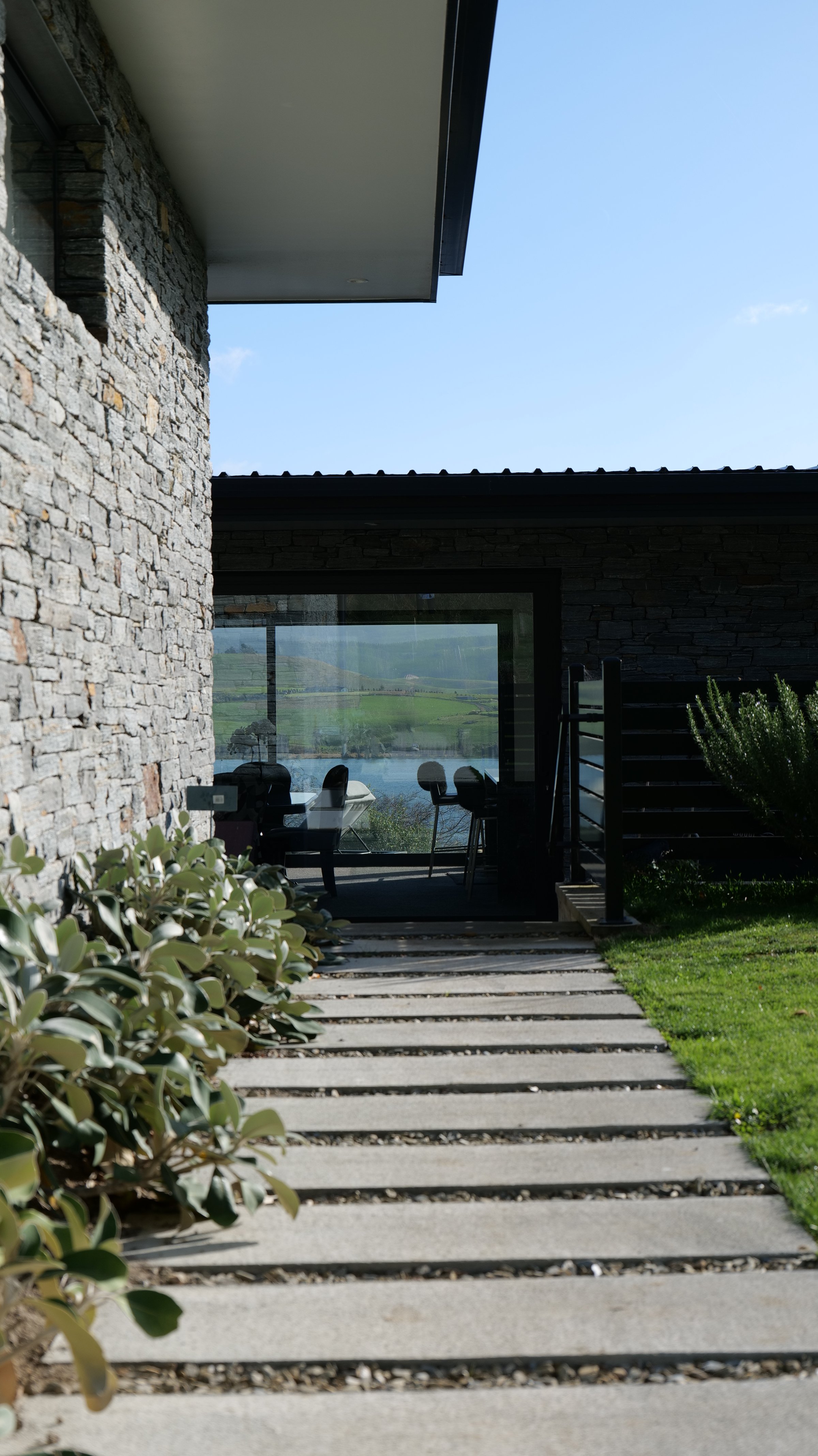 A modern house with stone exterior walls and large glass doors, viewing a scenic lake and green fields outside. There are outdoor chairs and greenery near the entrance.