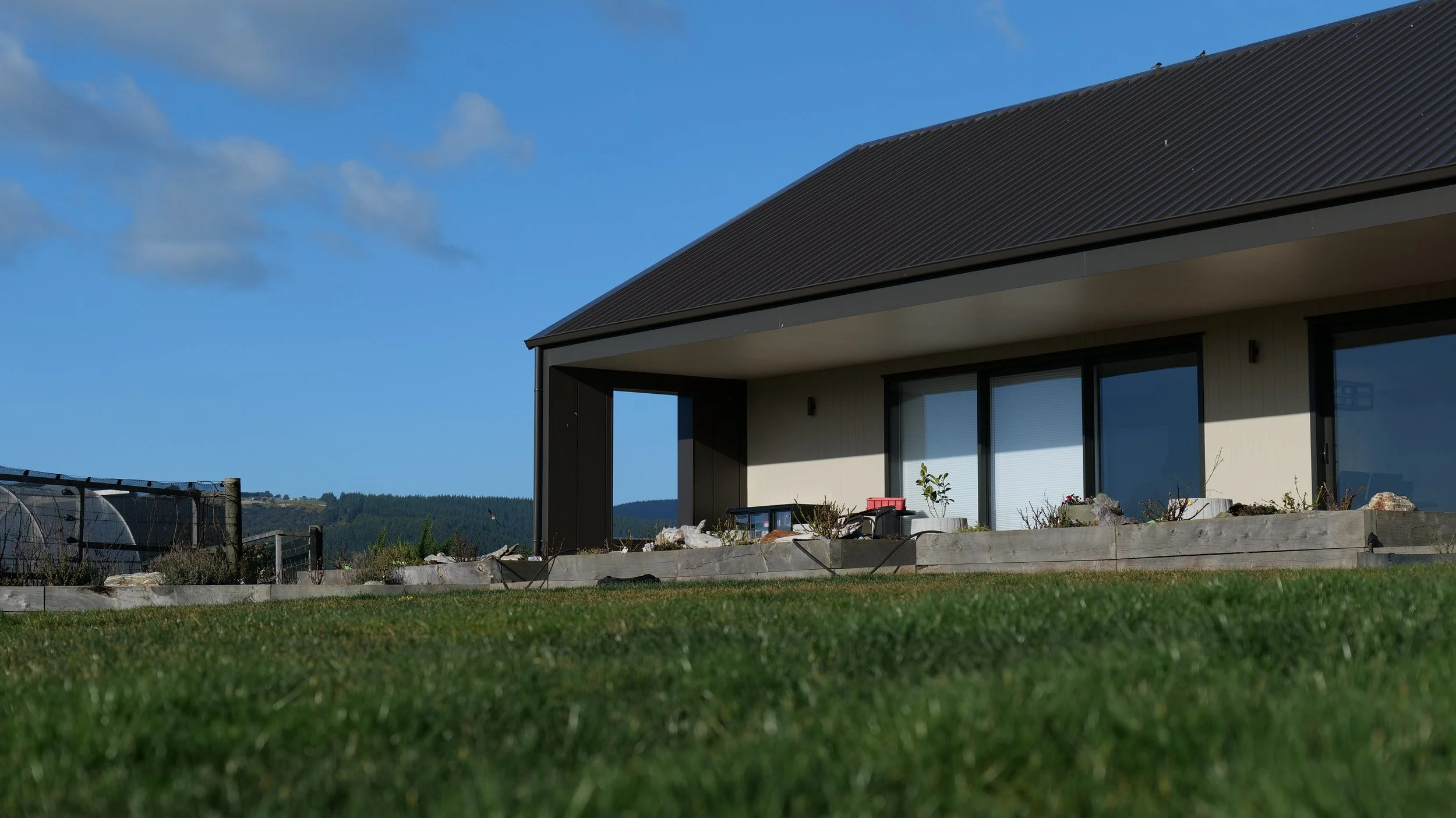 Modern house with large windows and a sloped roof under a blue sky, with a grassy yard and a stone border in the foreground.