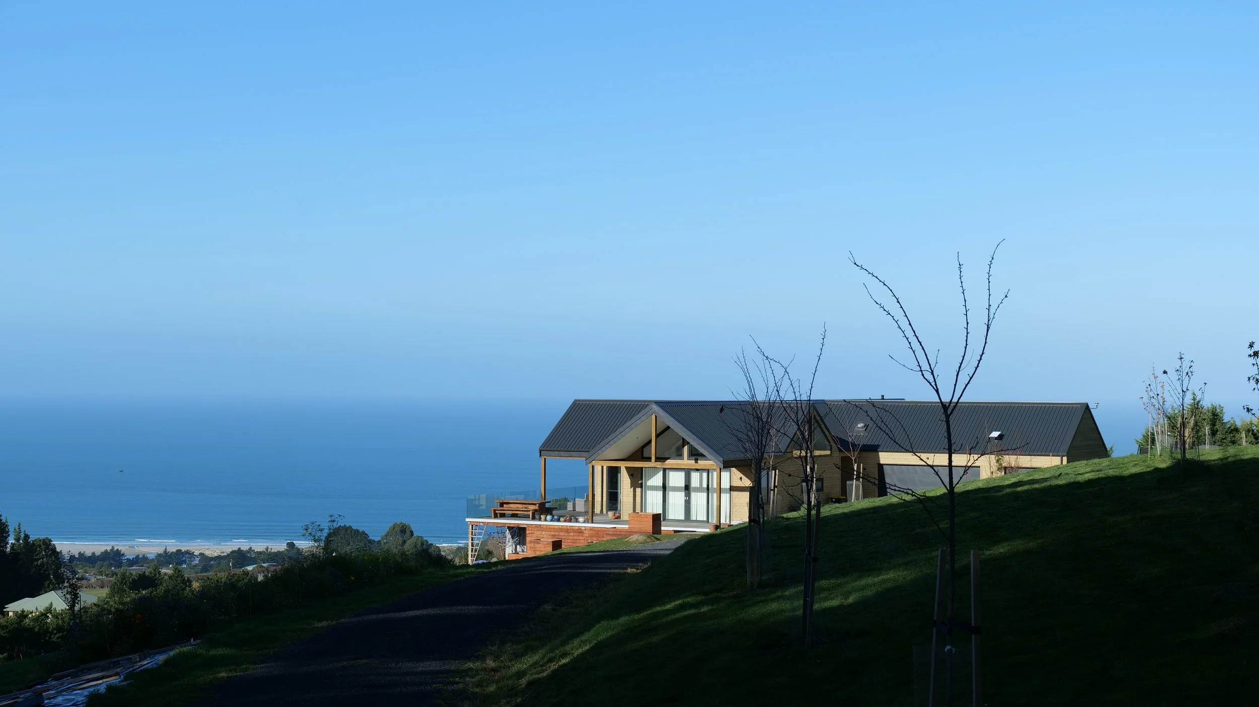 Modern house with large windows on a hill, overlooking a body of water, with trees and a clear blue sky.