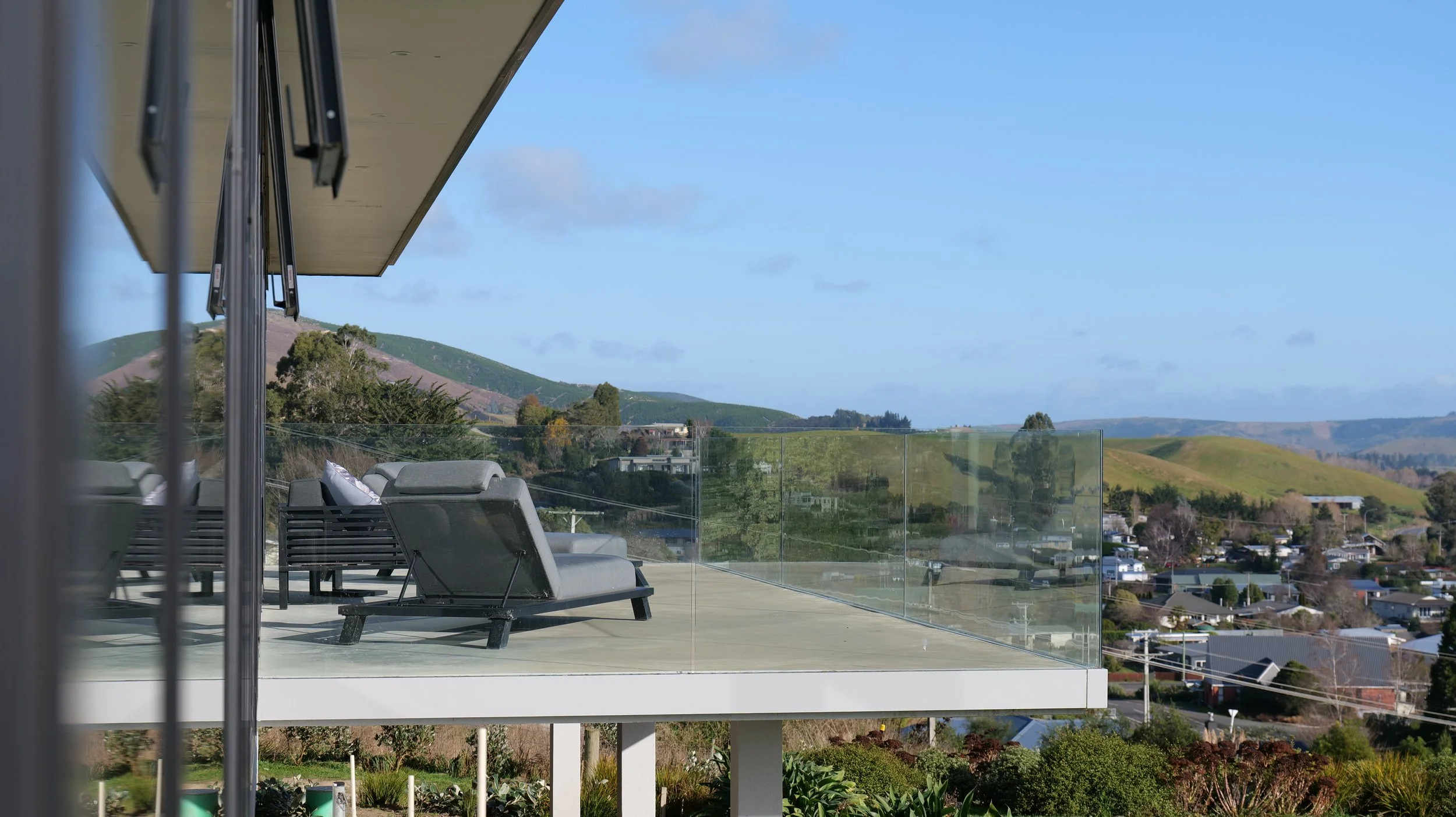 Balcony with lounge chairs and glass railing overlooking hills and residential area under blue sky