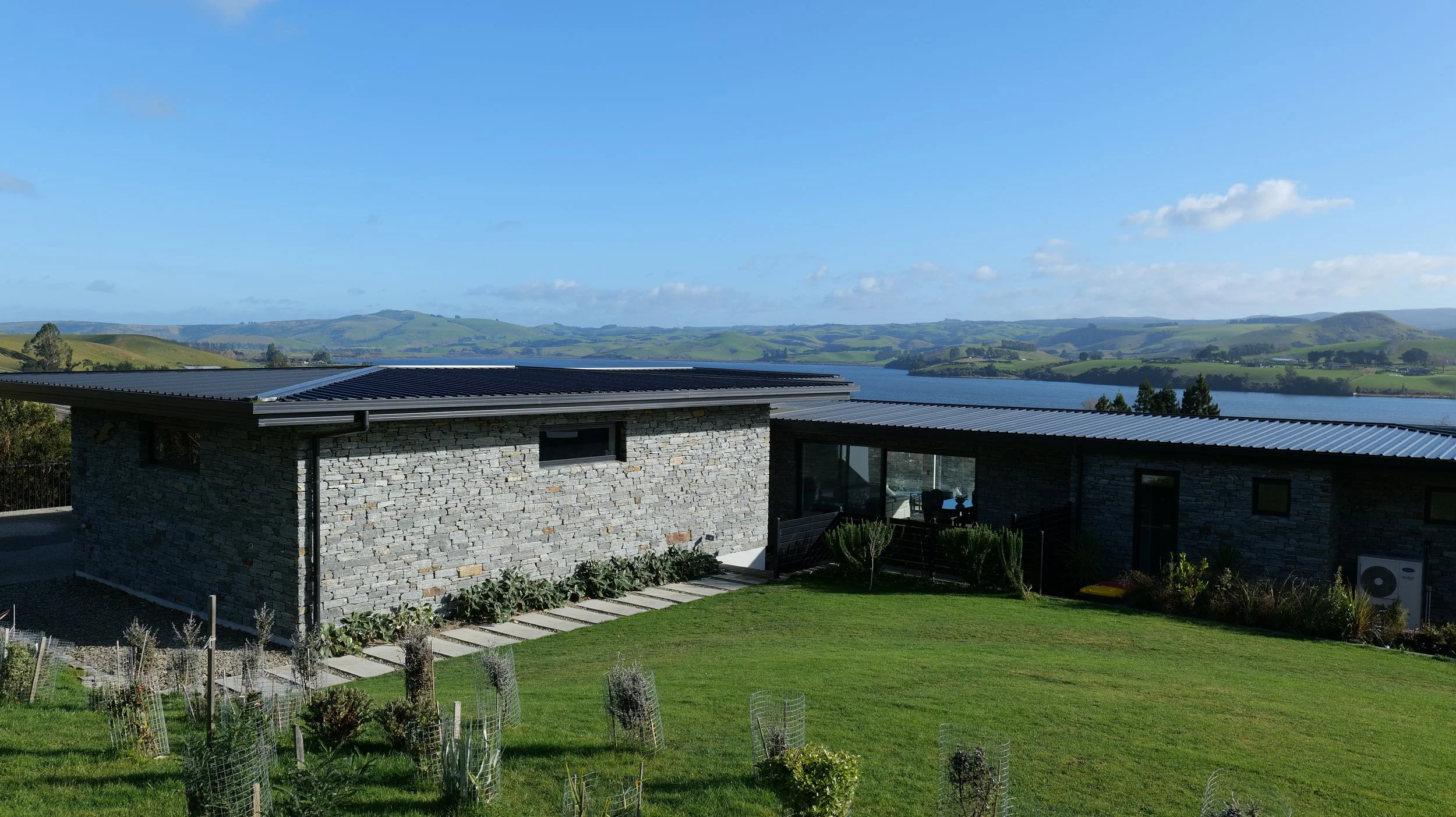 Modern stone house with large windows, green lawn with garden beds, overlooking a river and green rolling hills under a partly cloudy sky.