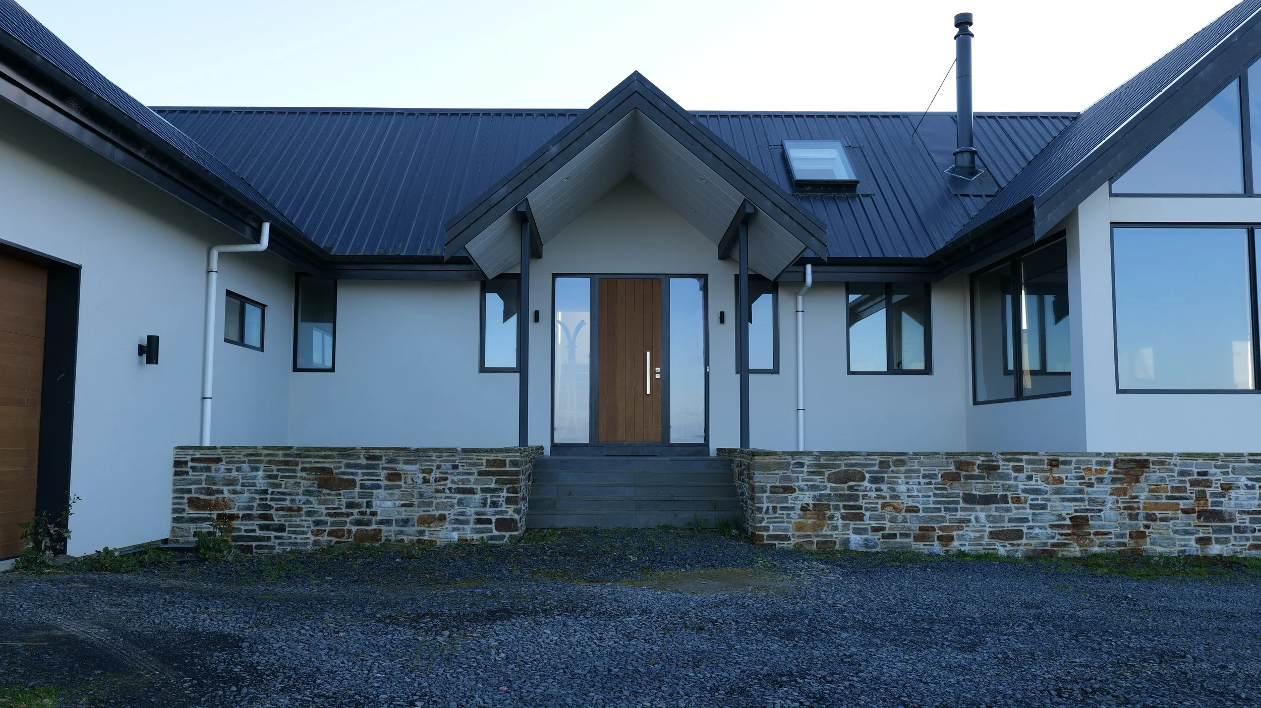 Front view of a modern house with a dark metal roof, white walls, stone foundation, and a wooden front door, with large windows.