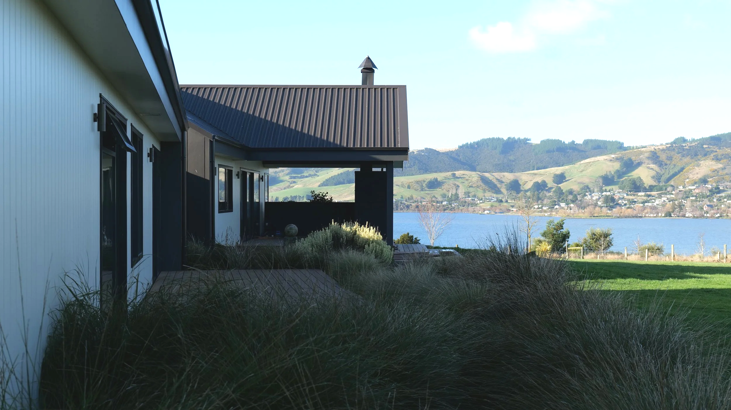 View of a lakeside house with a metal roof, surrounded by tall grass and plants, with water and hills in the background.