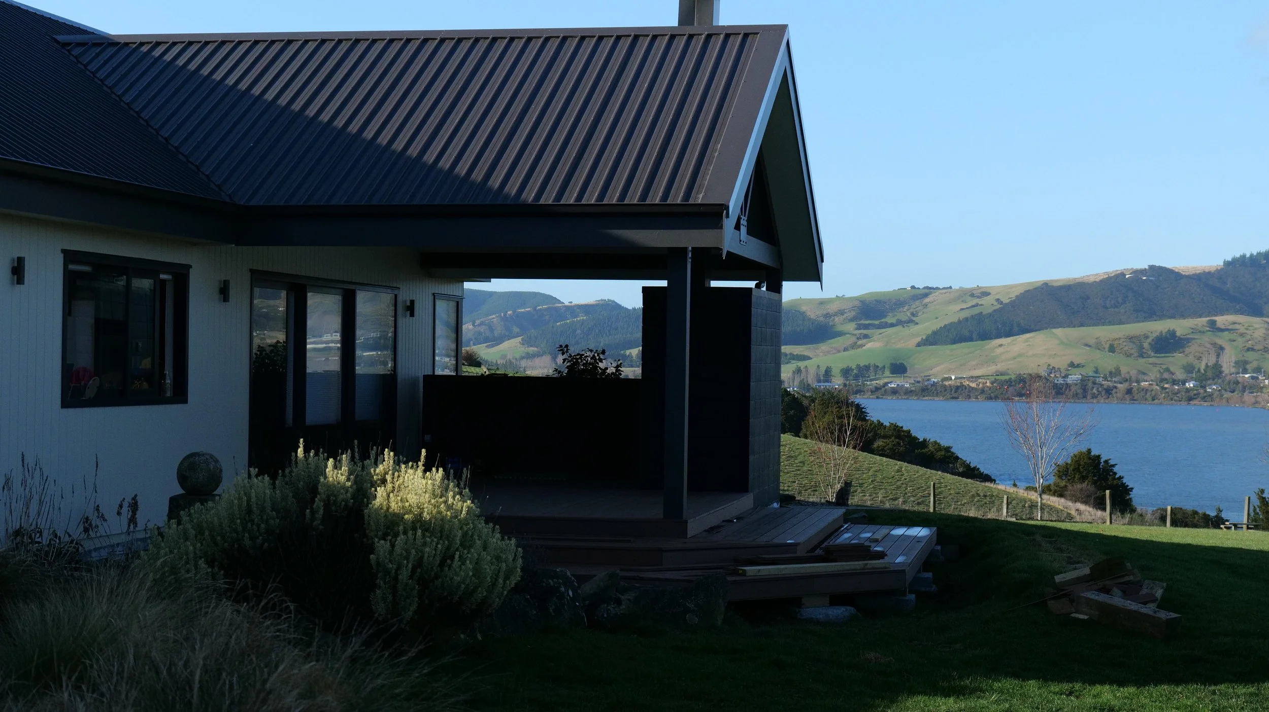 Modern house with a black metal roof, large windows, and a deck overlooking a lake with rolling green hills in the background.