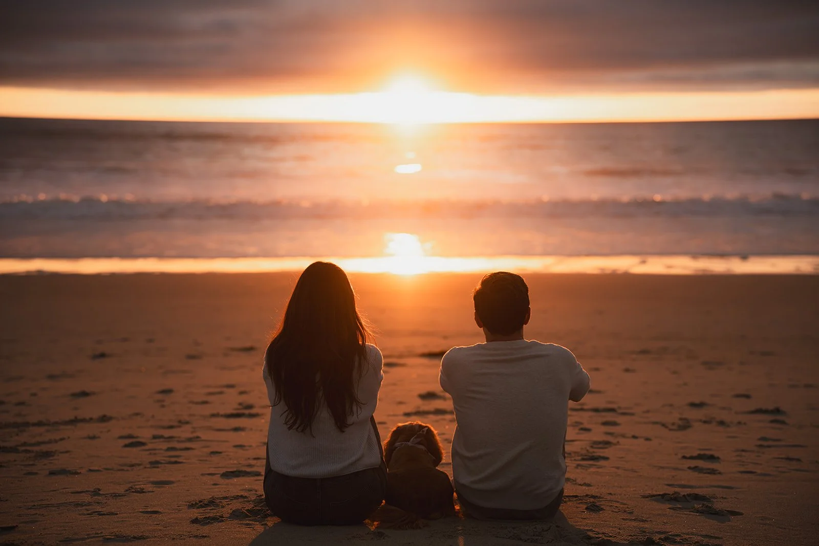 A couple and a dog sitting on the beach at sunset, watching the ocean and the sun setting over the horizon.
