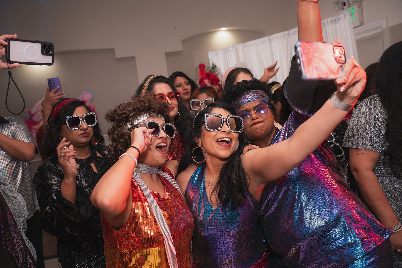 A group of women dressed in colorful, shiny outfits and large, decorative sunglasses taking a selfie at a party.