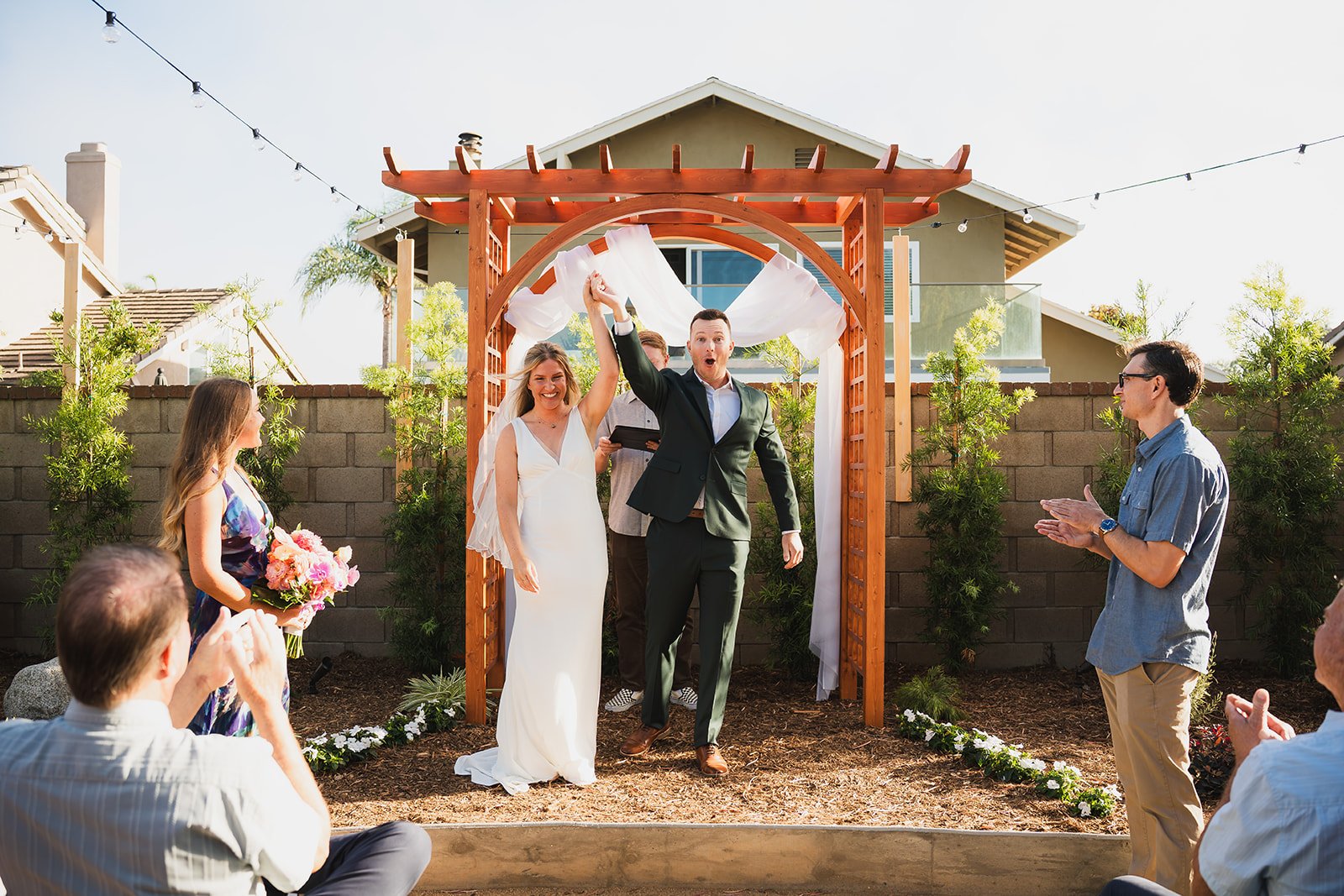 A couple is celebrating their wedding in an outdoor ceremony, with the bride in a white gown, the groom in a suit, and guests clapping and watching under a wooden arch decorated with white fabric.