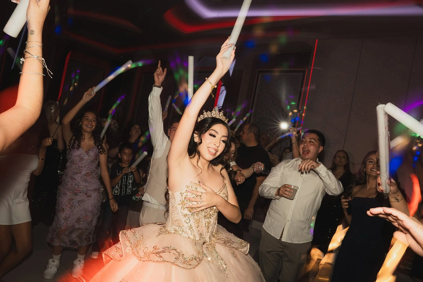A young woman in a peach-colored dress and tiara dances joyfully at a celebration, surrounded by smiling guests holding glow sticks in a dimly lit room with colorful light streaks.