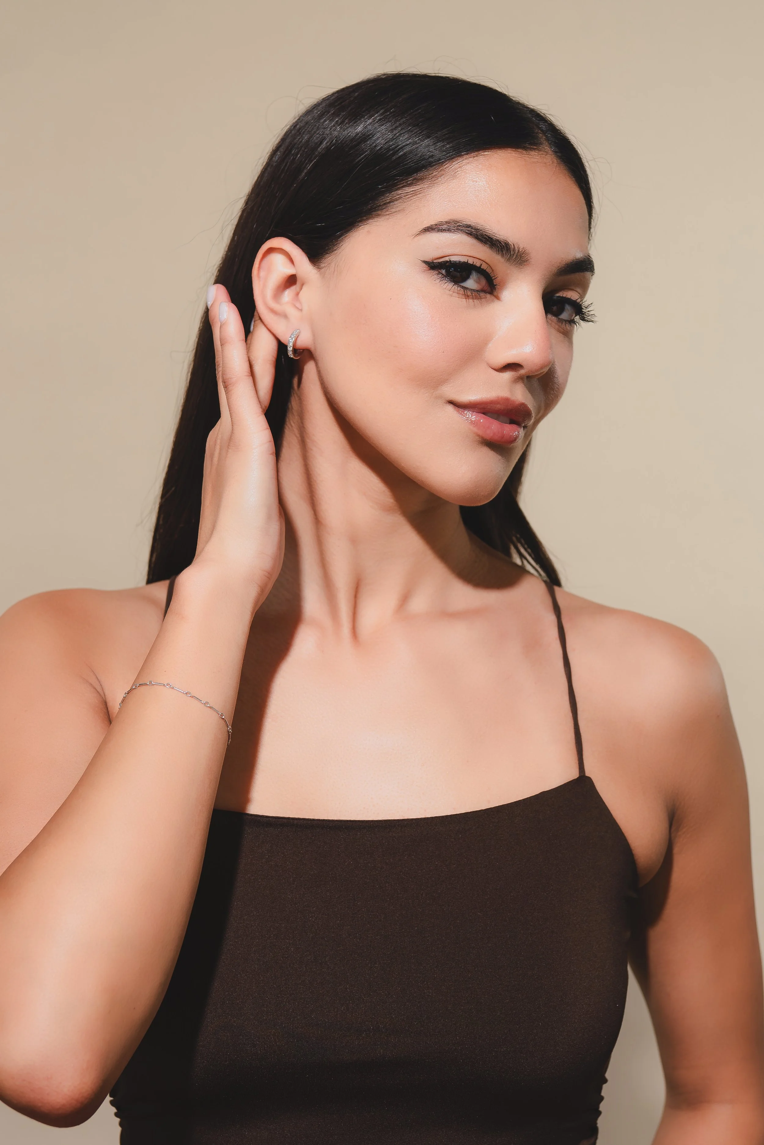 A woman with long dark hair, wearing a black spaghetti strap top, poses with her right hand touching her ear, looking at the camera with a slight smile.