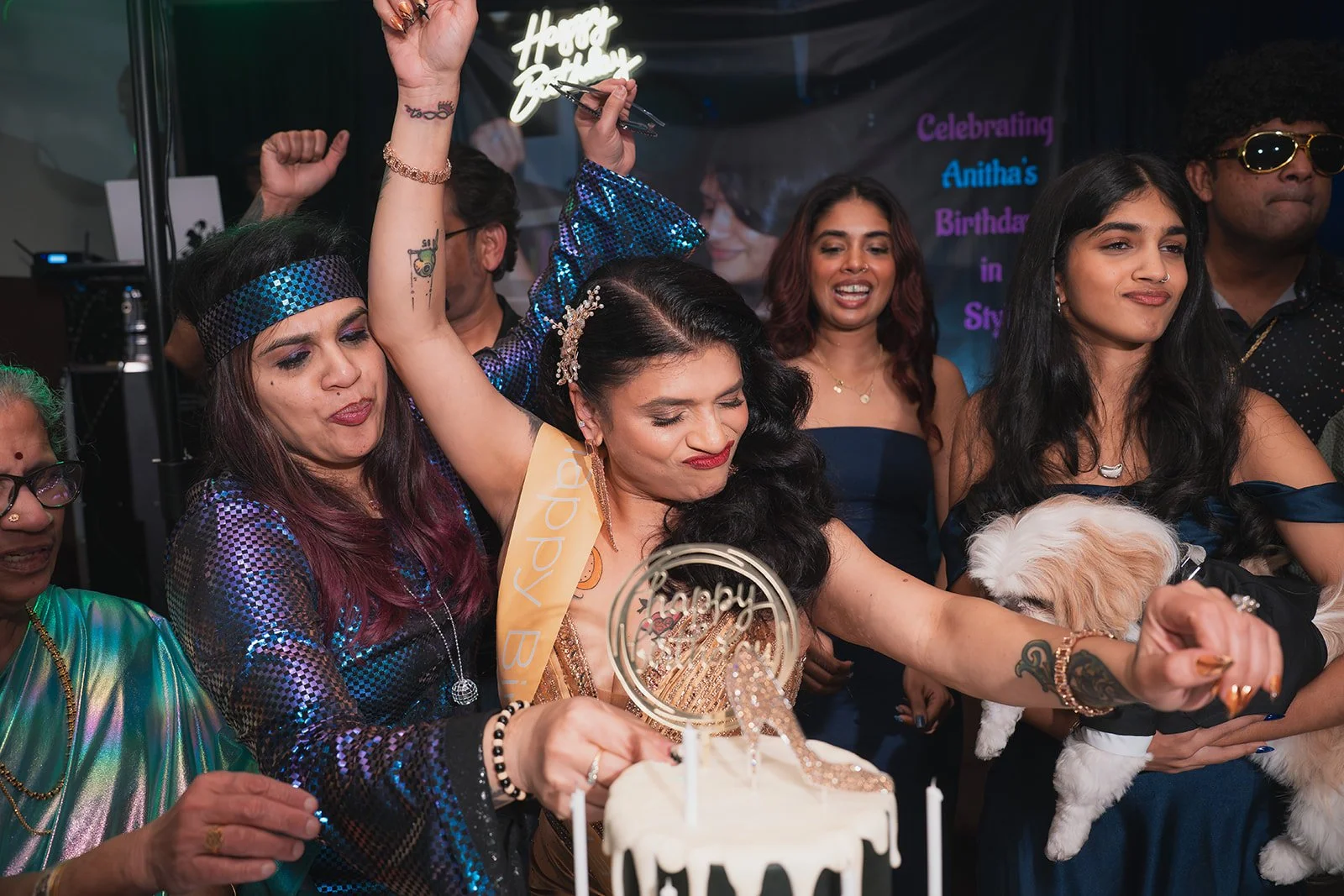 Women celebrating a birthday, with one woman wearing a sash, surrounded by friends holding a dog, with a birthday cake in front, and a festive background with colorful writing.