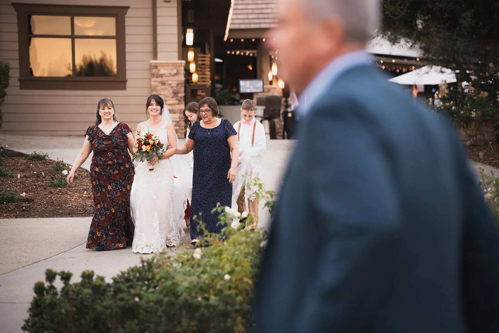 A bride walking with her family outside a building, followed by her groom and flower girl, while an older man in the foreground looks on.