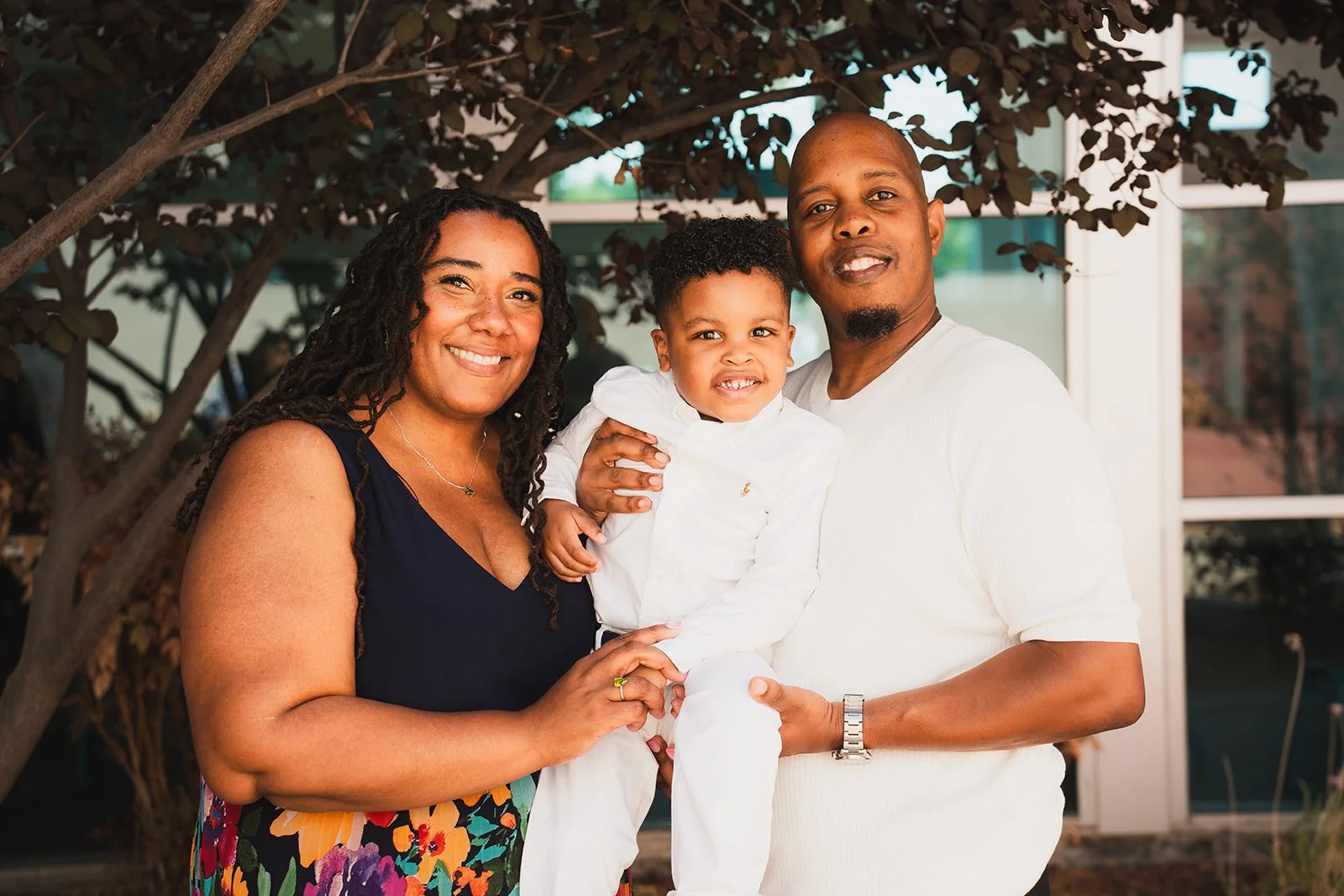 A family of three, including a woman with long dark curly hair wearing a dark top and floral skirt, a young boy in white clothes, and a man with close-cropped hair and a white shirt, standing outdoors in front of a tree and glass building.