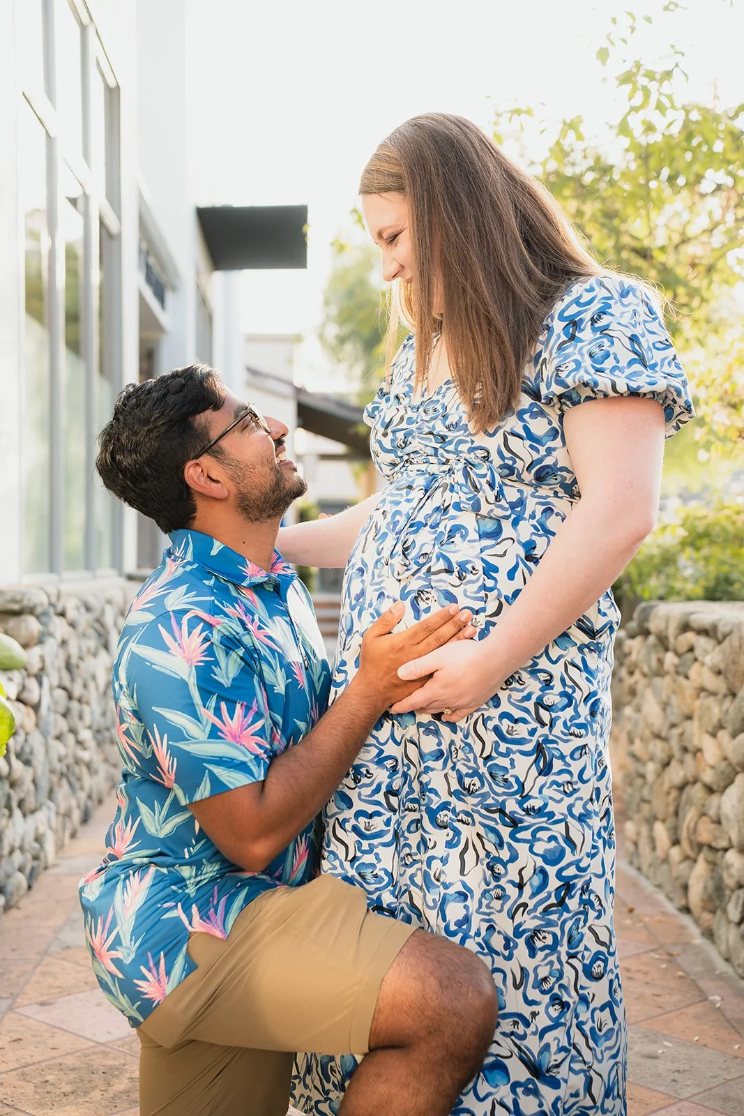 A man kneeling and holding the pregnant belly of a woman, smiling at each other outdoors with sunlight coming through trees.