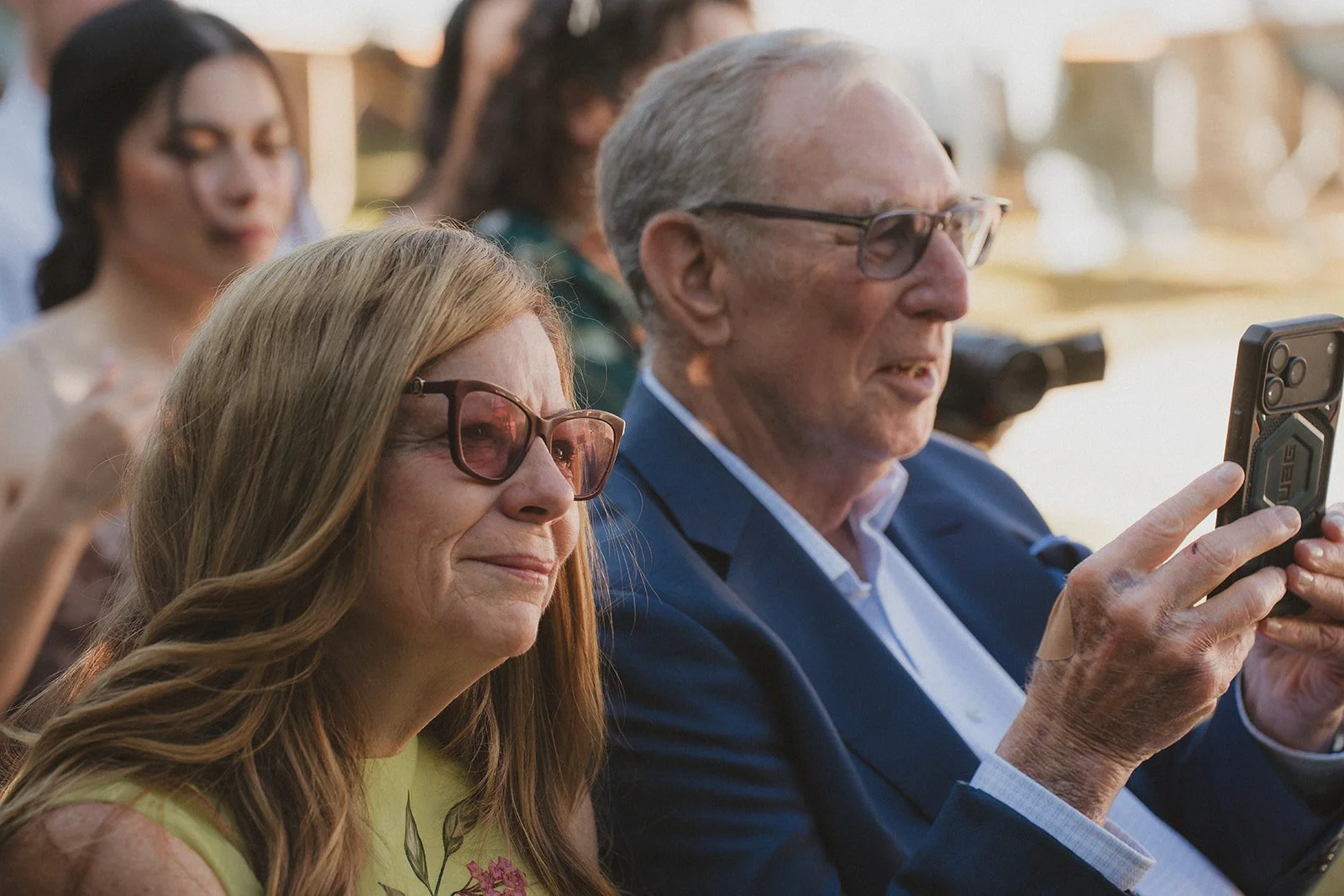 An elderly man and woman sitting outdoors, with the man holding a smartphone and the woman looking on, while others are in the background.
