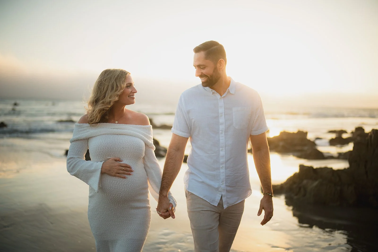 A pregnant woman and a man holding hands walking on the beach during sunset, smiling at each other.