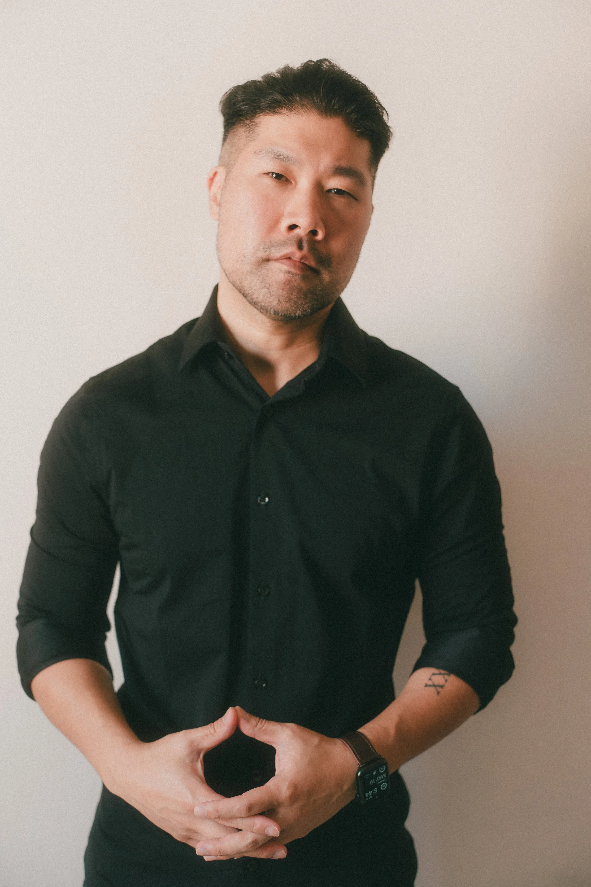 A man with dark hair, wearing a black shirt, standing against a plain light-colored wall