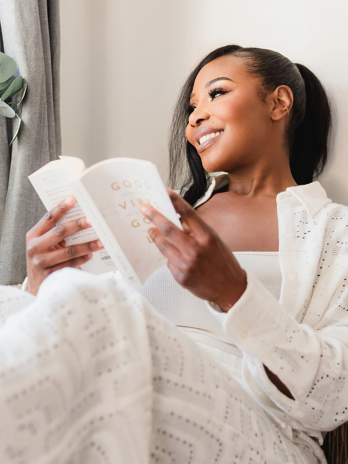 A smiling woman with dark hair in a ponytail, wearing a white outfit, is reading a book while sitting near a window with a curtain and a plant nearby.