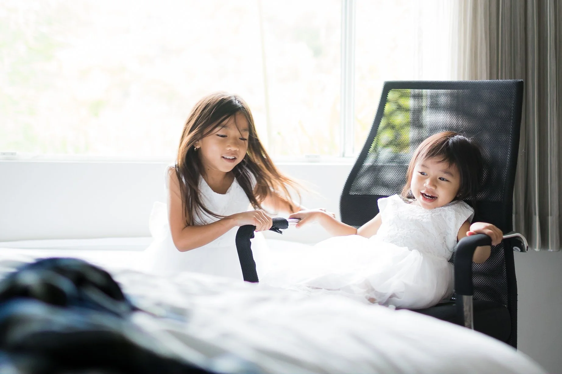 Two young girls in white dresses playing together on a bed. One girl is sitting in a black office chair, while the other girl is standing and helping her. They are in a bright room with large windows.