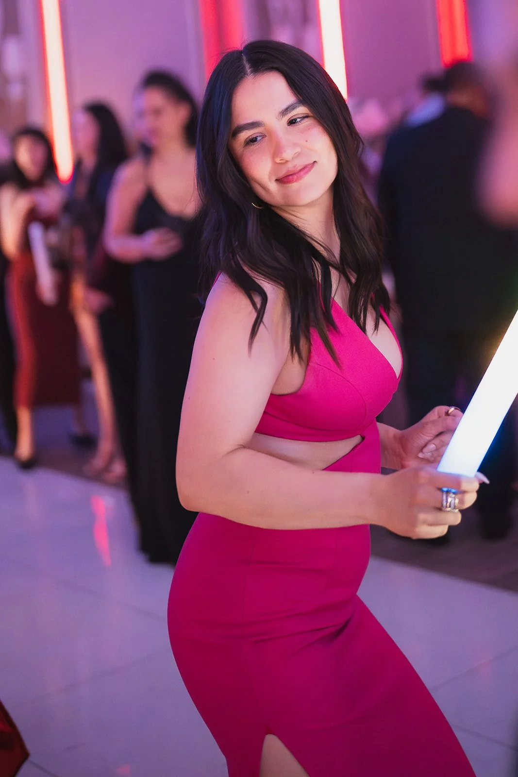 A young woman with dark hair wearing a pink dress, holding a lighted panel, smiling at a party or social event.