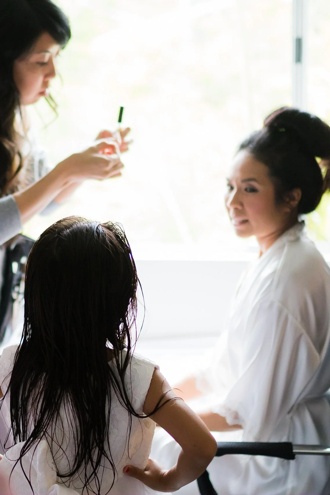 Women preparing a bride for her wedding by fixing her hair and makeup, with a young girl in an ivory dress watching.