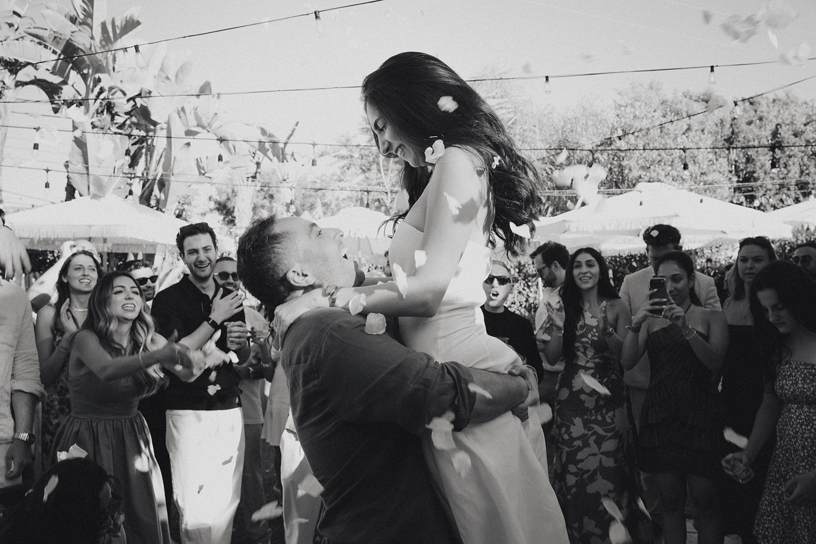 A man lifts a woman in a dress with friends watching and smiling at an outdoor celebration with umbrellas and string lights.