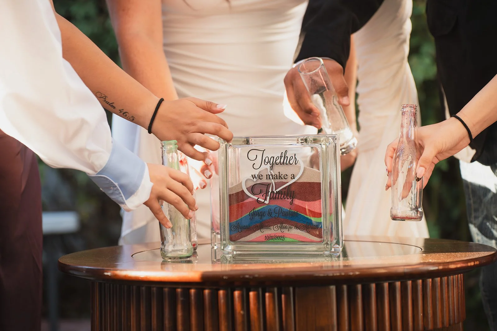People holding empty glasses around a glass box with a rainbow design and the text "Together we make a Family" on it, indicating a wedding or family event.