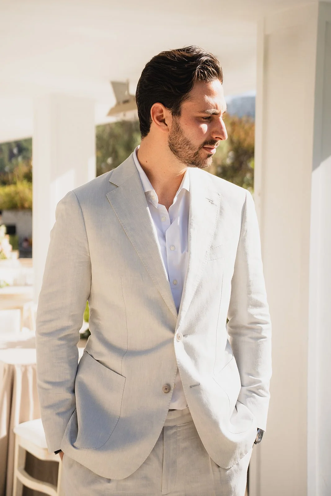 A man in a white suit and shirt standing indoors near a window, looking to the side.