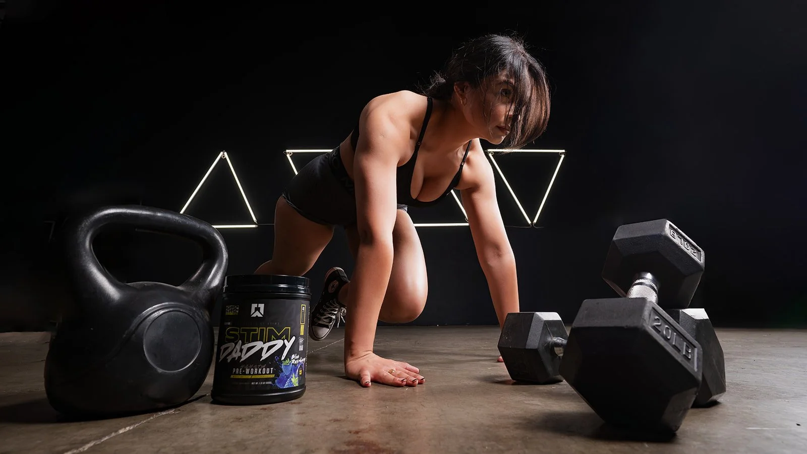 A woman in workout clothes on the floor in a gym with kettlebell, dumbbells, and pre-workout supplement container.