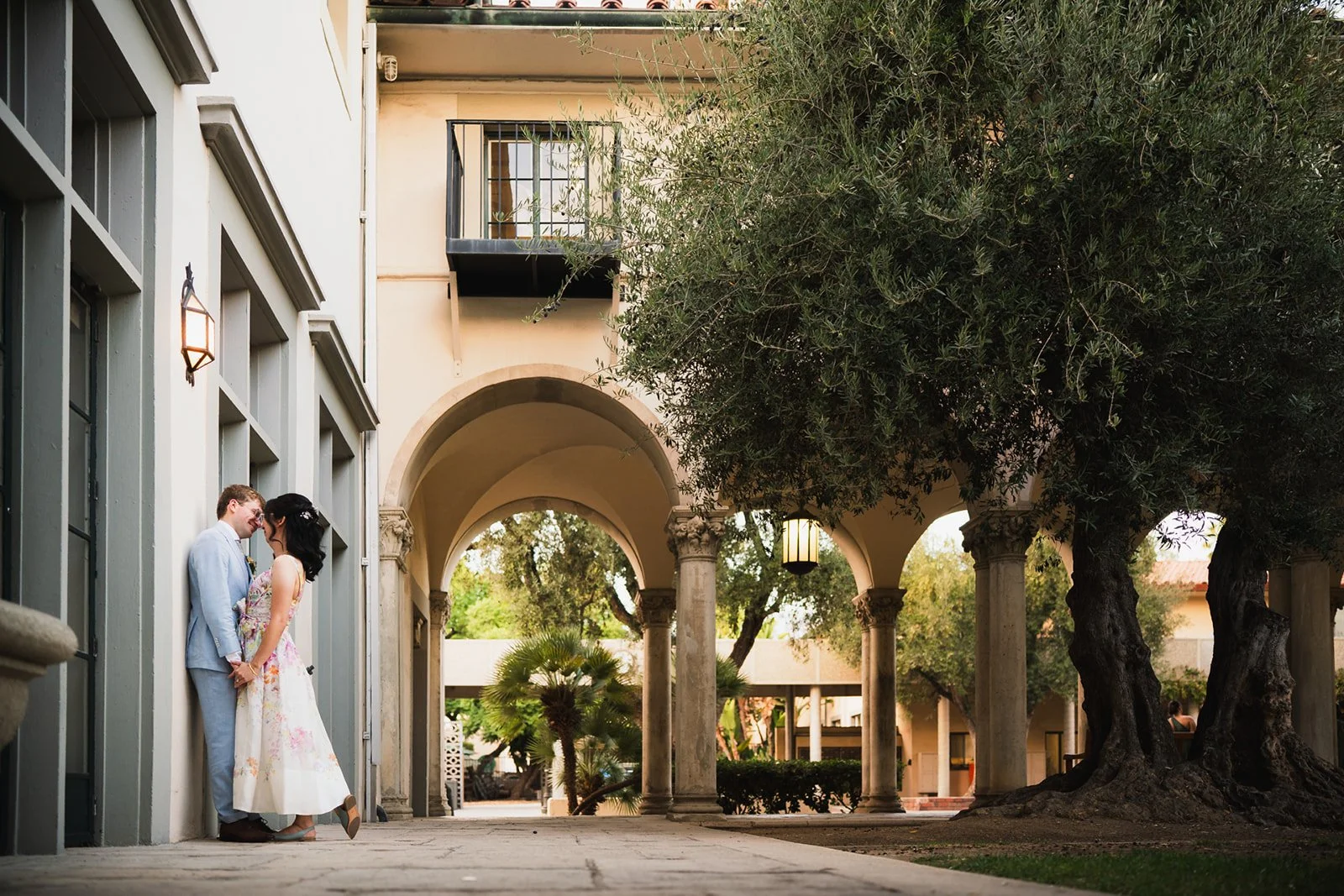 A couple dressed in wedding attire sharing a moment under an archway surrounded by trees and colonial-style columns.