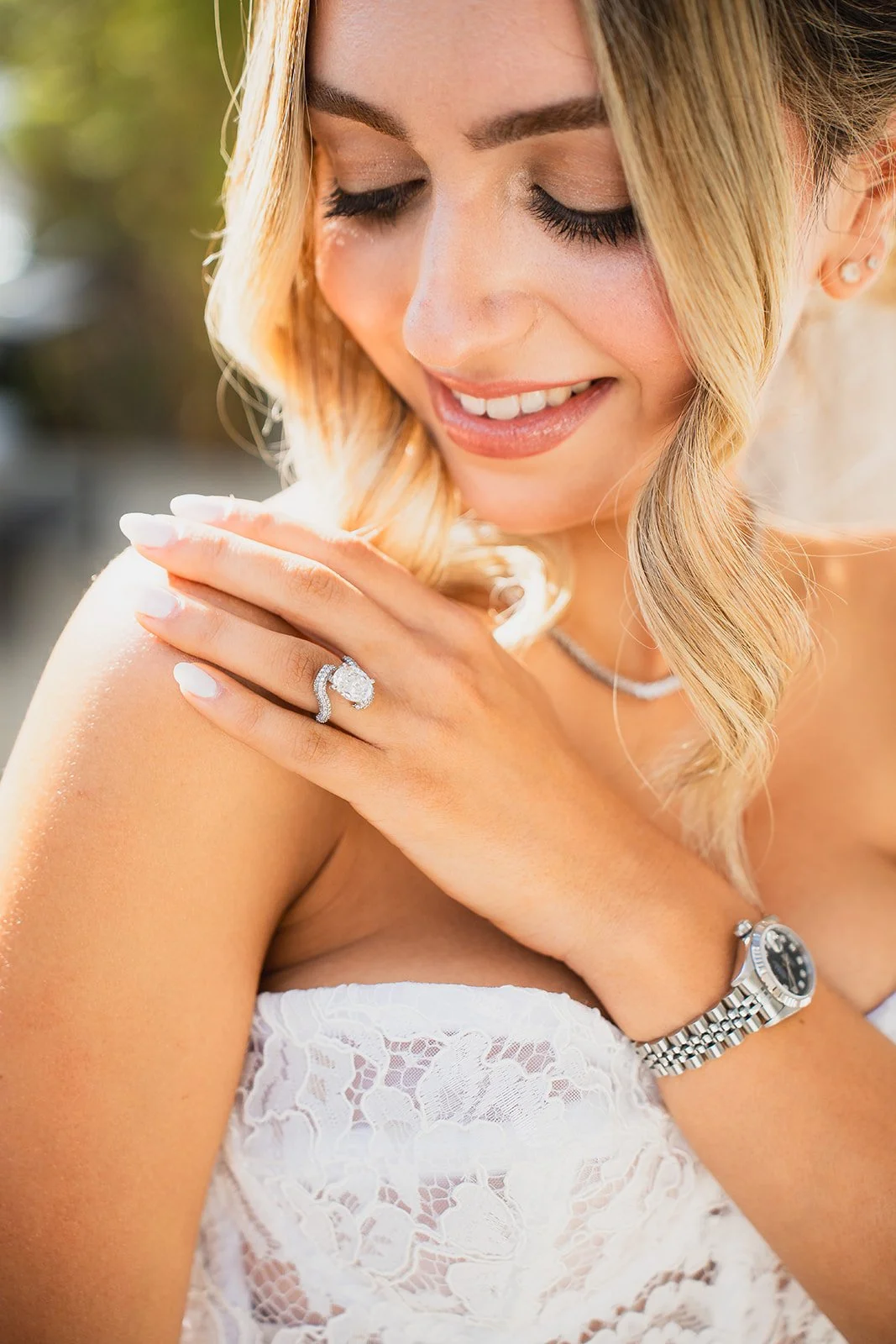 Close-up of a woman with blonde hair, smiling with eyes closed, showing off a diamond engagement ring, a watch, and jewelry on her neck and ear.