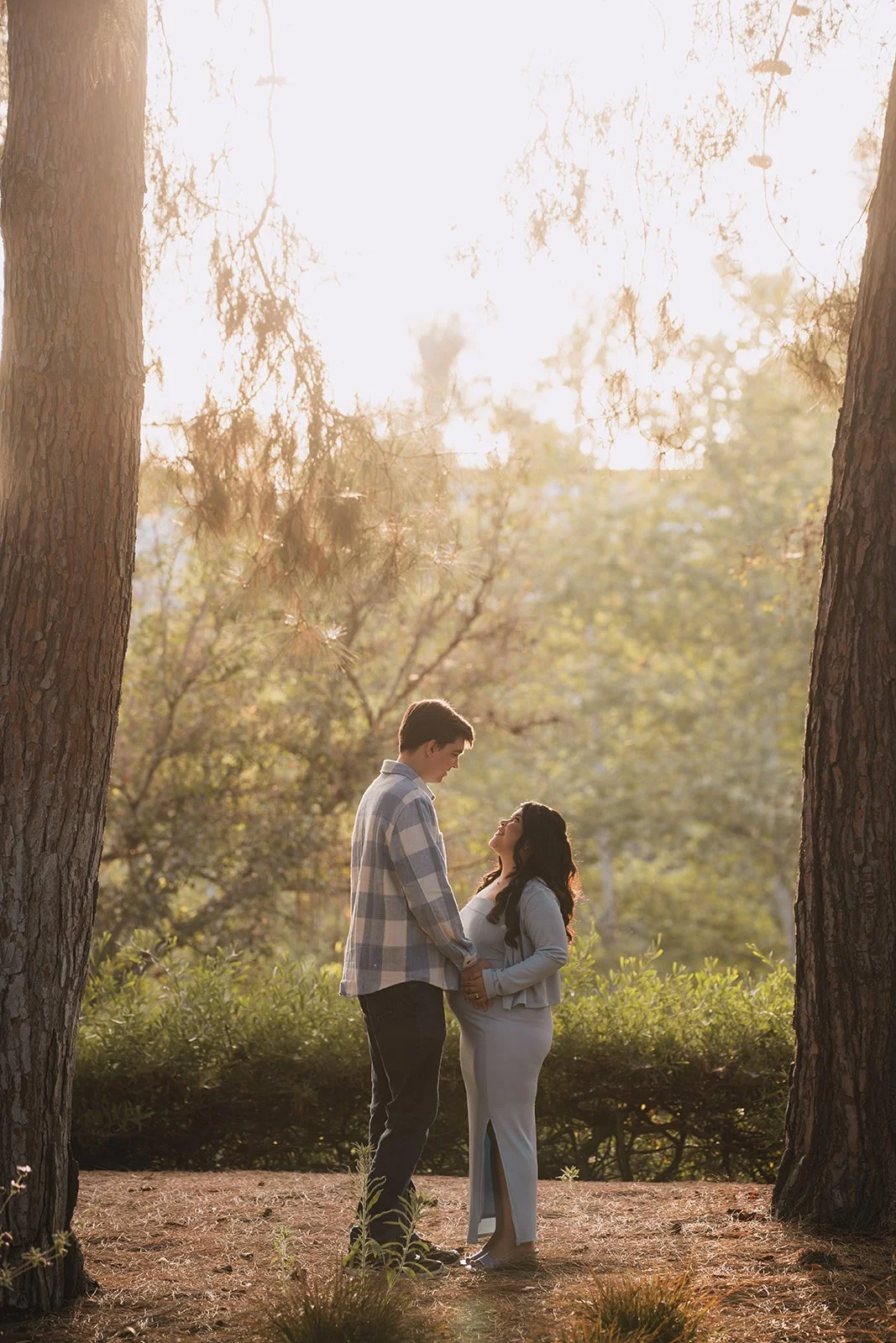 A couple holding hands and gazing at each other in a forest with sunlight streaming through trees.