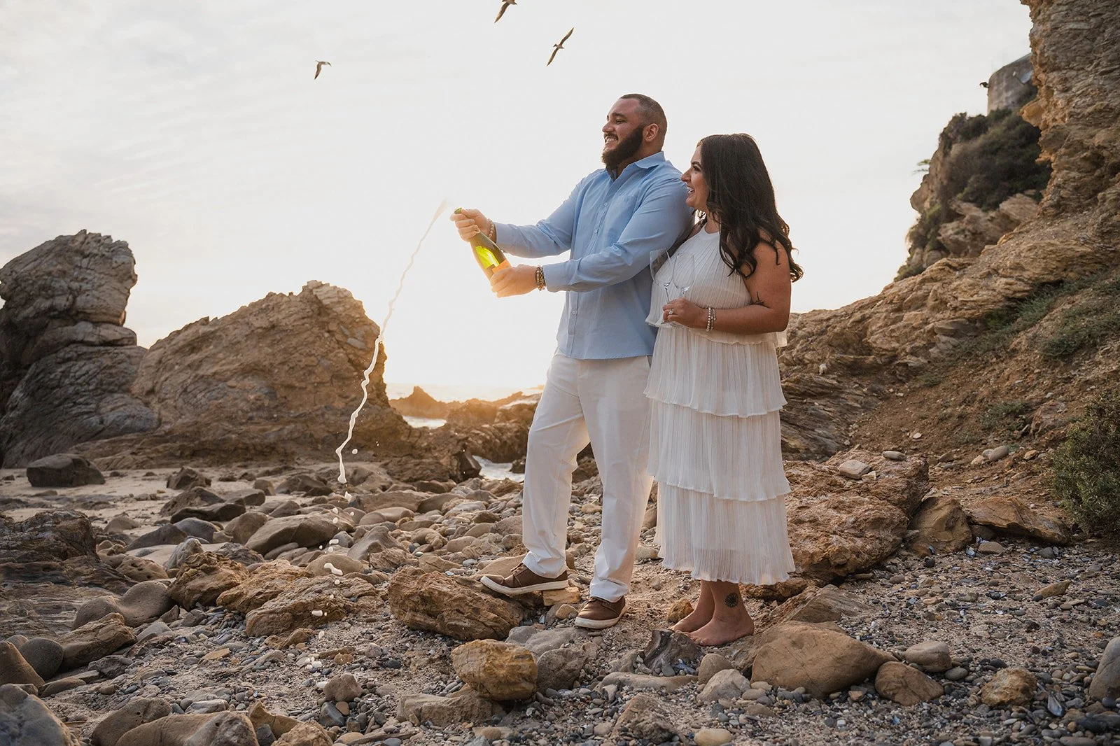 A couple celebrating on a rocky beach with a bottle of champagne, the man opening it and the woman holding a glass, during sunset.